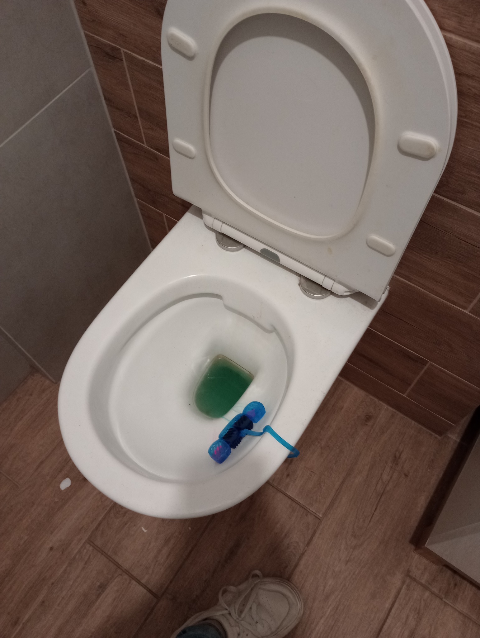 Biała toaleta z otwartą klapą, kostka toaletowa w muszli, brązowe płytki podłogowe i szare ścienne w tle. Widok z góry.