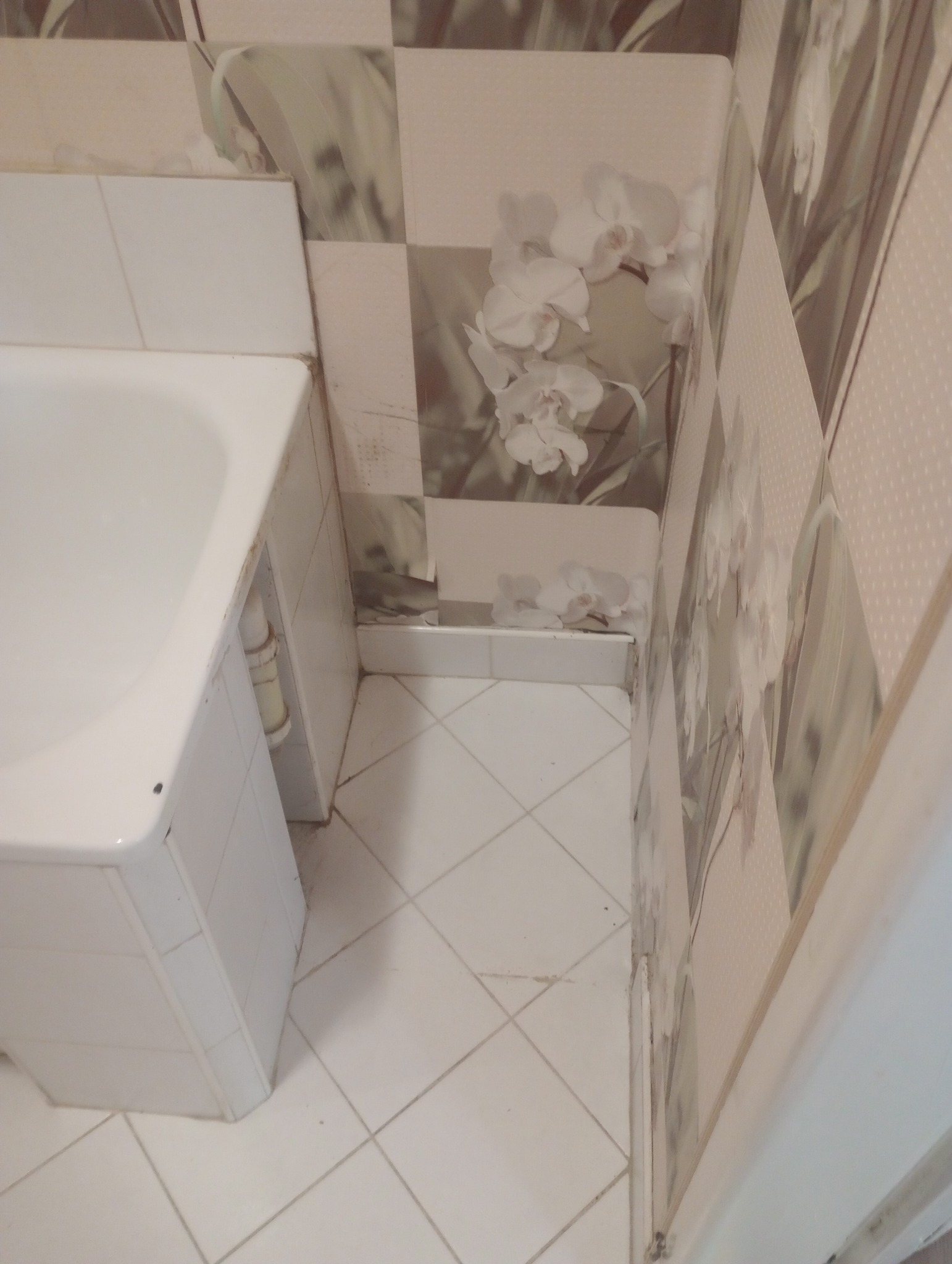 Fragment łazienki z białą wanną, jasnymi płytkami podłogowymi i dekoracyjnymi panelami ściennymi z motywem kwiatowym, ukazujący stan wymagający interwencji sprzątającej.