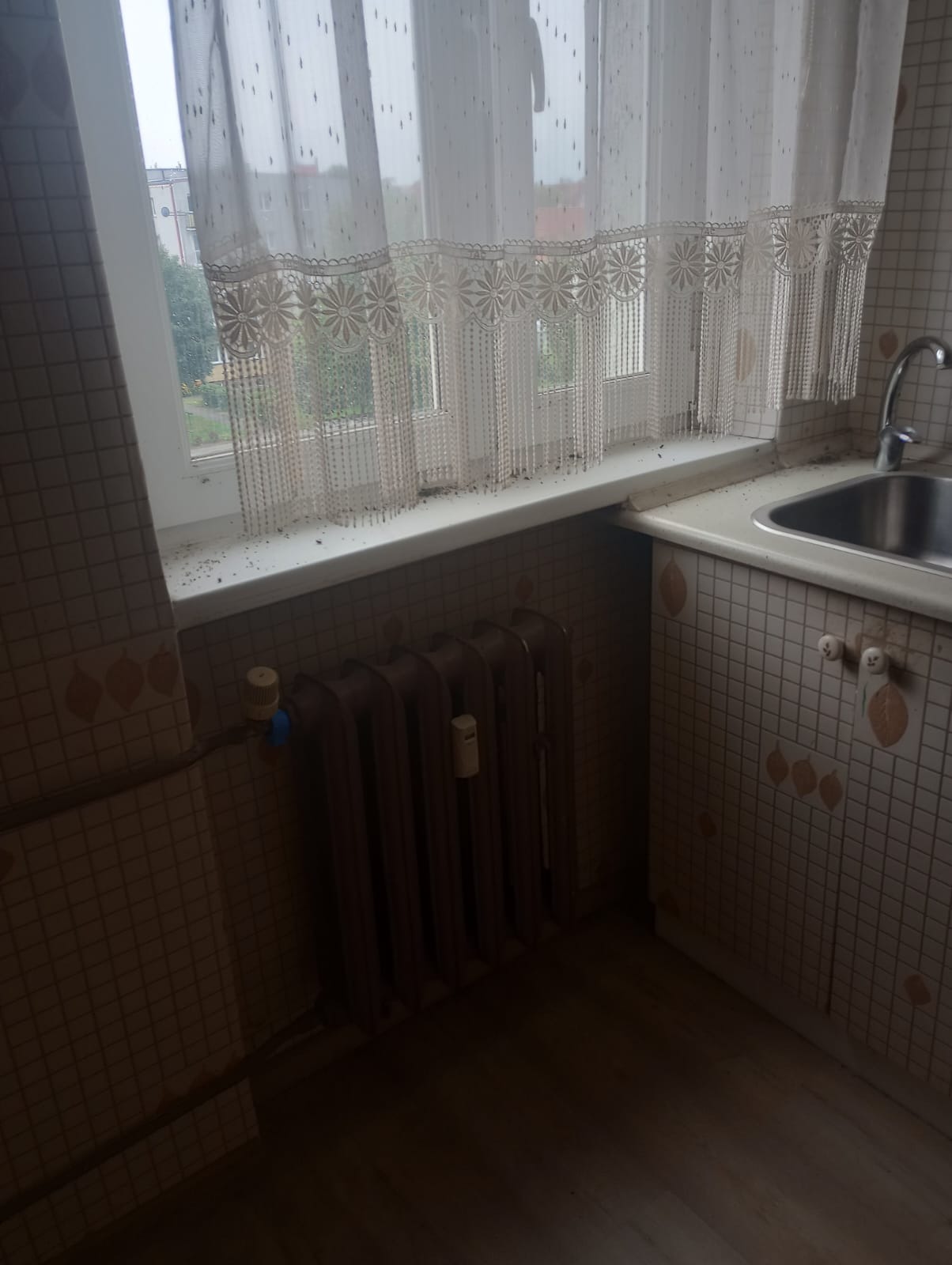 Zaniedbana kuchnia z brudnym oknem, zlewem i grzejnikiem. Widoczne zabrudzenia na parapecie, firance i podłodze. Potrzebne sprzątanie.