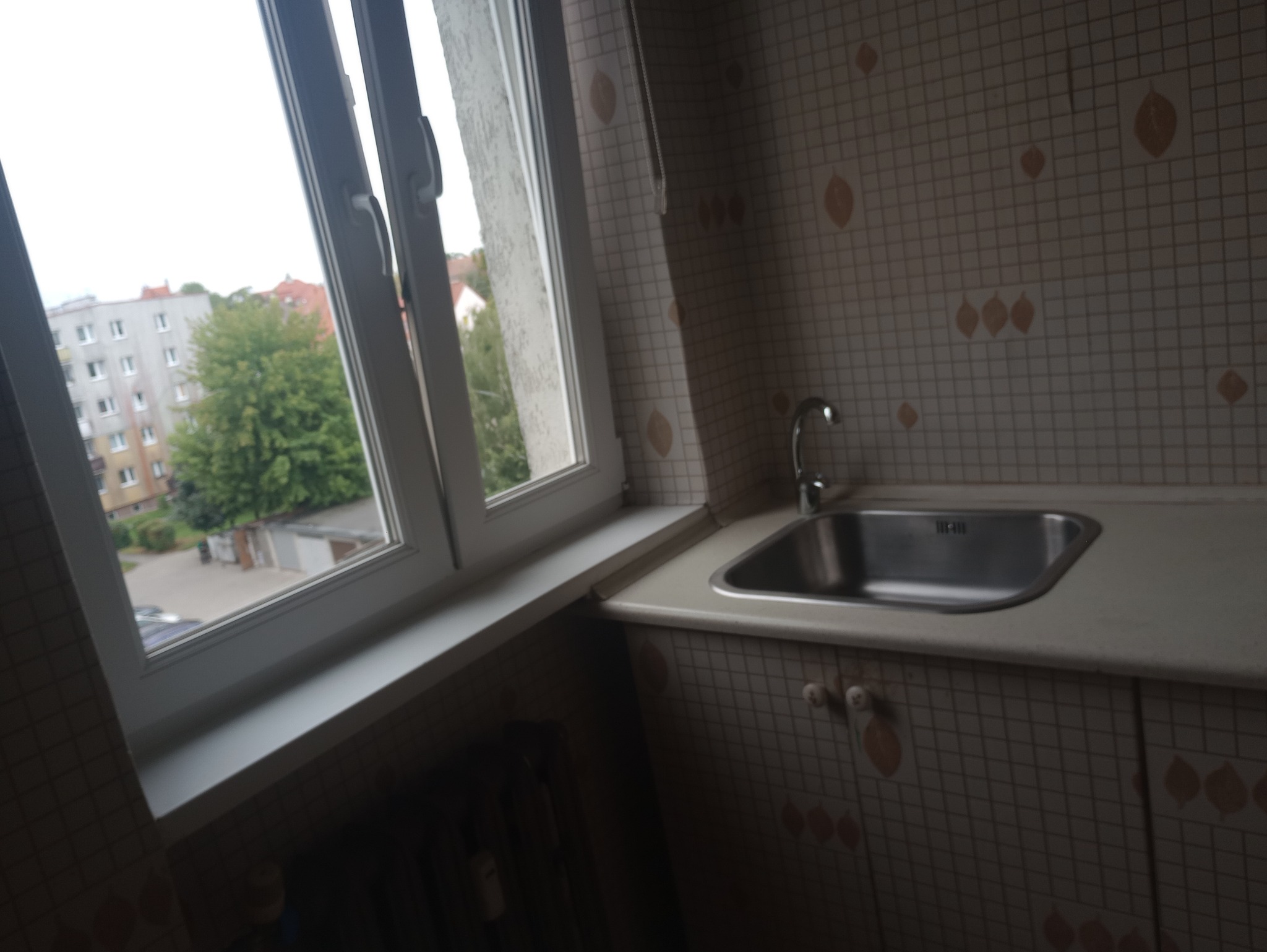Umywalka w kuchni z widokiem na blok przez okno. Beżowe kafelki z motywem liści. Metalowa bateria i zlew. Widok na zieleń i zabudowania za oknem.
