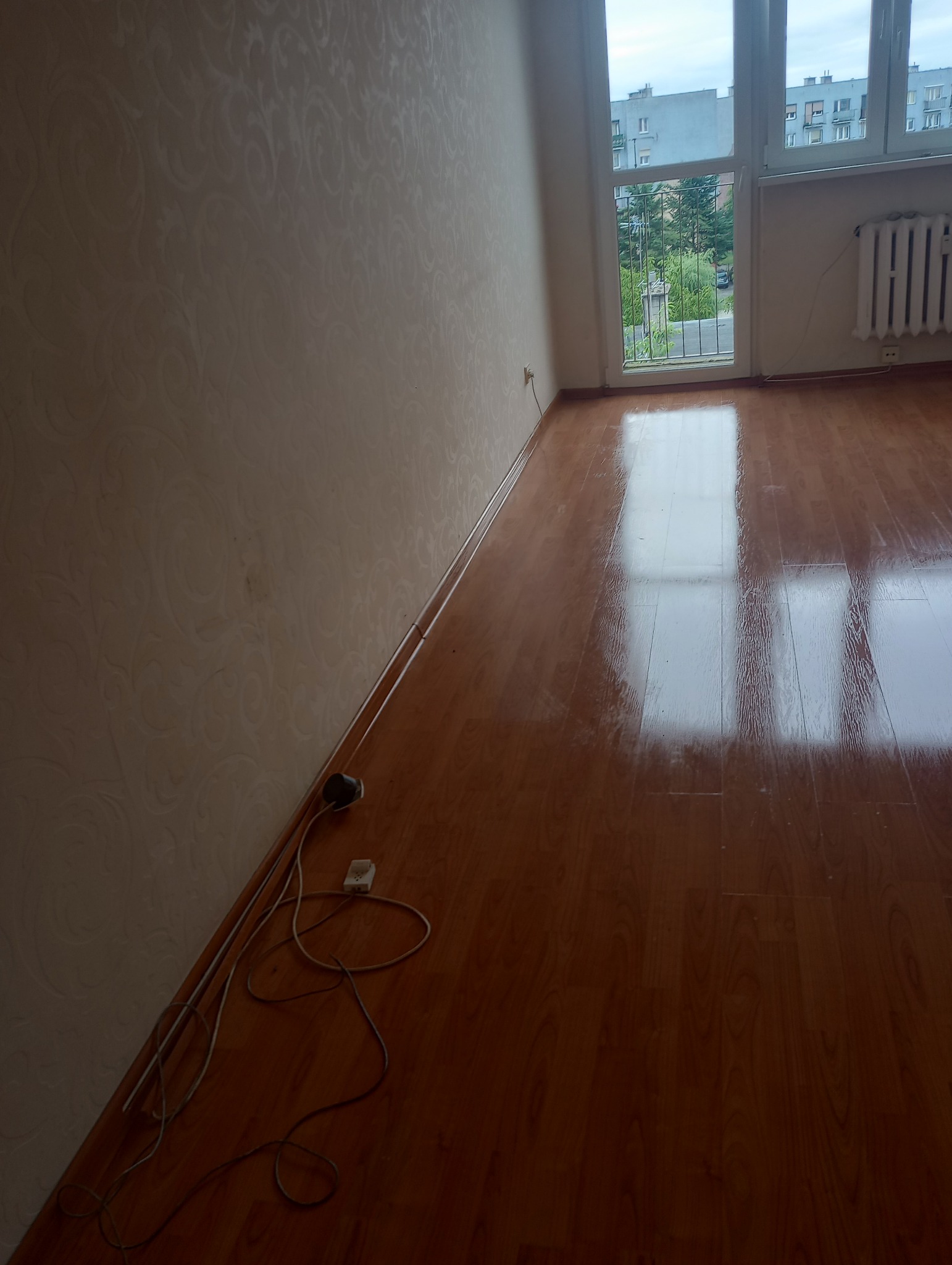 Wnętrze pokoju po remoncie: świeżo umyta podłoga z paneli odbija światło z okna. Tapeta w delikatny wzór, widoczne gniazdko i kable przy ścianie.