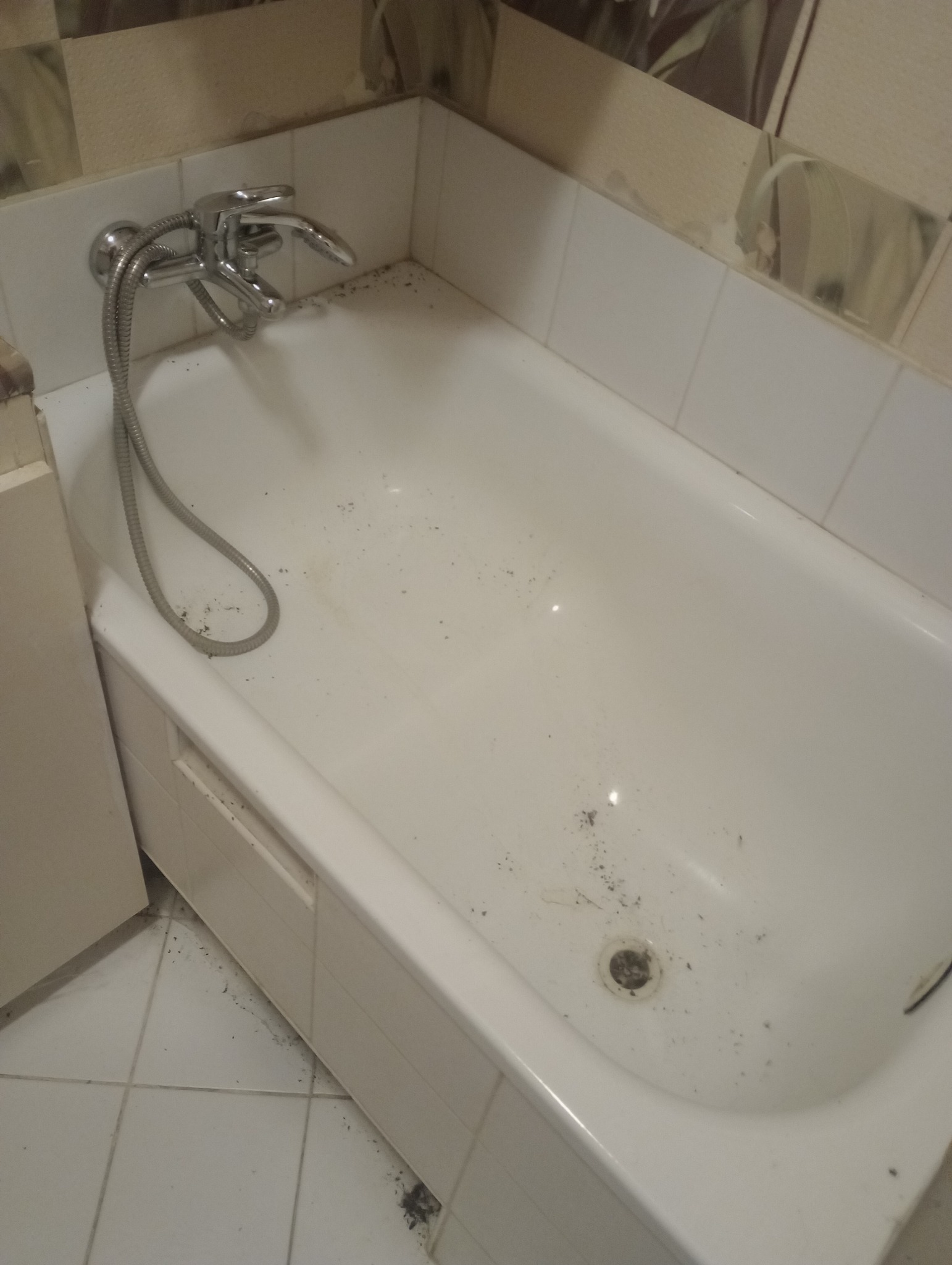 Brudna, biała wanna z chromowaną armaturą, widoczne zabrudzenia na powierzchni wanny i wokół odpływu, płytki na ścianach i podłodze, wnętrze łazienki przed sprzątaniem.