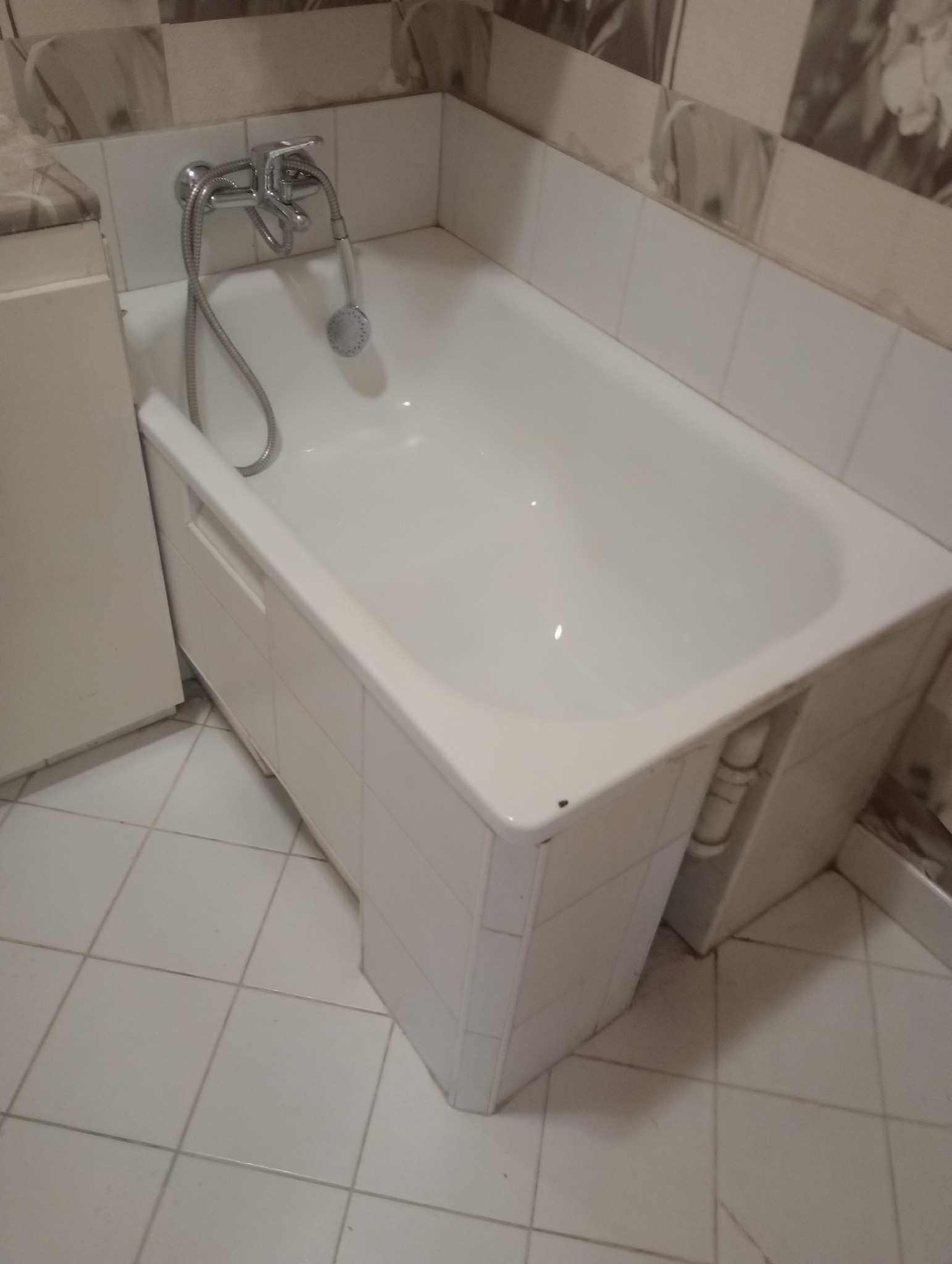 Biała wanna z baterią i słuchawką prysznicową w łazience z białymi kafelkami. Widok z góry na czystą wannę i fragment podłogi z jasnych płytek.