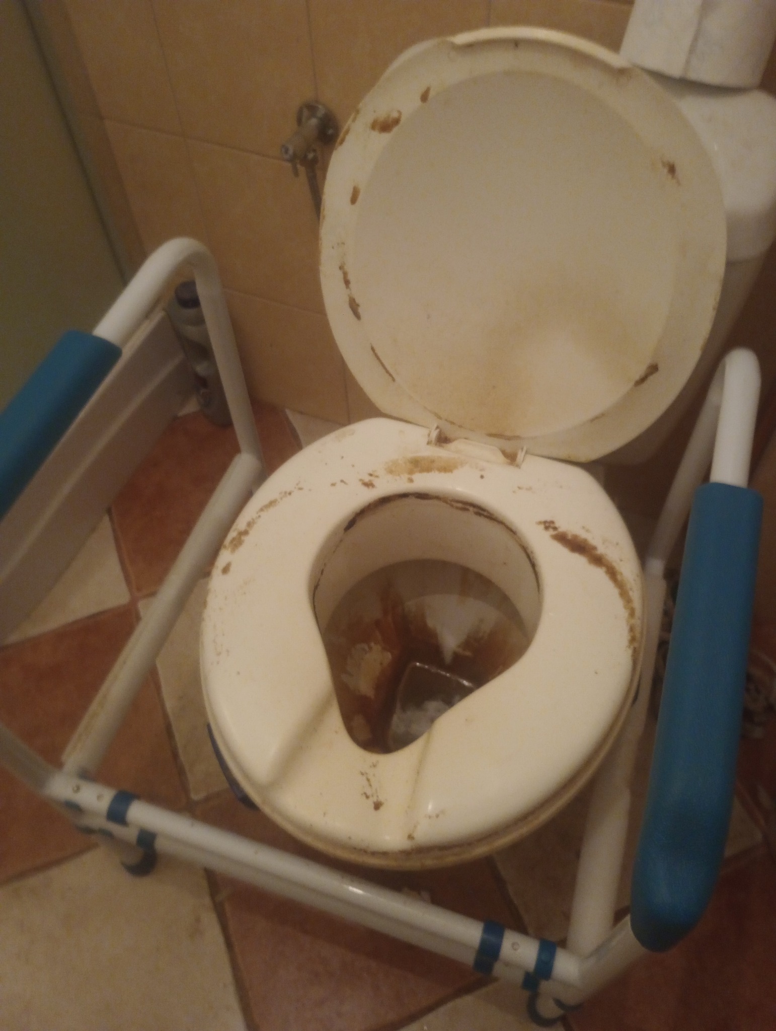 Z bliska: bardzo zabrudniona toaleta z podwyższeniem i uchwytami, widoczne brązowe zacieki i osady. Ujęcie z góry, słabe oświetlenie.