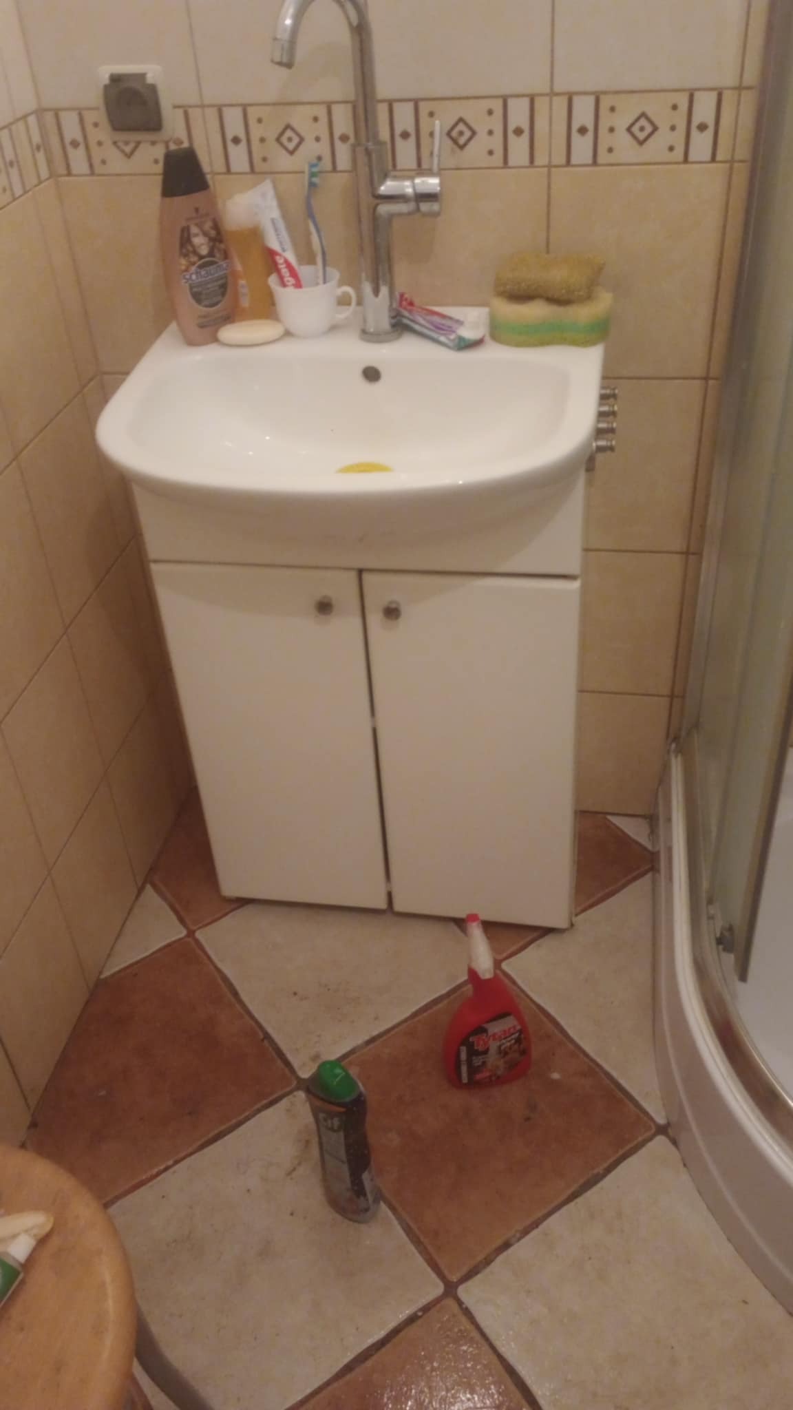 Umywalka z szafką w łazience po sprzątaniu, widoczne środki czystości na podłodze. Beżowe płytki na ścianach i brązowo-beżowe na podłodze.