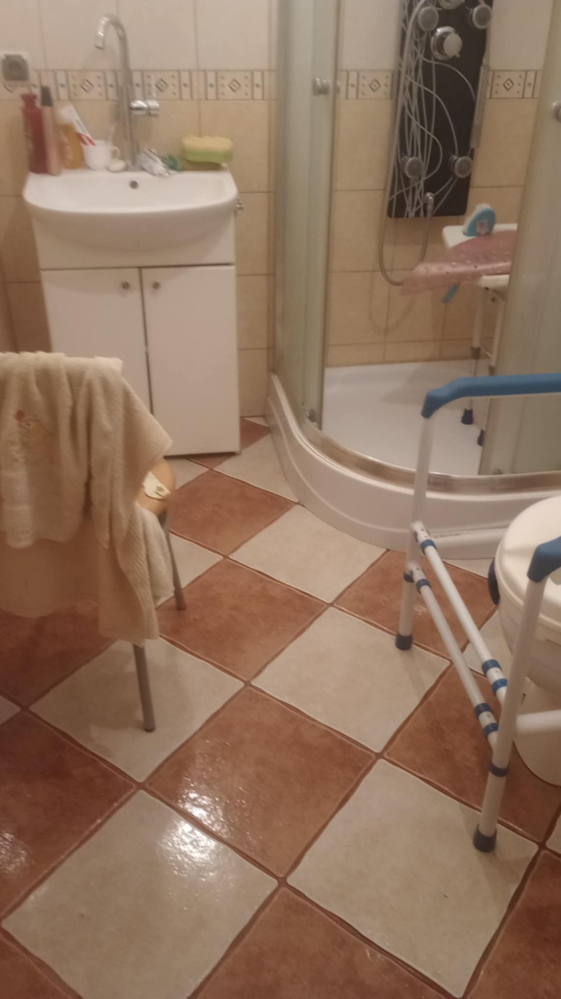 Czysta łazienka z brązowo-białymi płytkami, kabiną prysznicową, umywalką z szafką i krzesłem z ręcznikiem. Widoczny również sprzęt ułatwiający korzystanie z toalety.