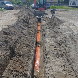 Elektro IT Sp.z.o.o - Wykop z pomarańczową folią ochronną w ziemi, przygotowany pod kable. W tle minikoparka i ubijarka wibracyjna. Widok z góry na plac budowy.