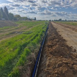 Elektro IT Sp.z.o.o - Błękitne rury kablowe ułożone w wykopie ziemnym na tle zielonej trawy i błękitnego nieba z chmurami. Instalacja kablowa w terenie otwartym.