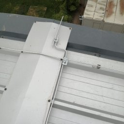 Elektro IT Sp.z.o.o - Metalowa instalacja odgromowa na dachu budynku z blachy trapezowej, widok z góry. Elementy połączone śrubami, tło stanowią kontenery i zieleń.