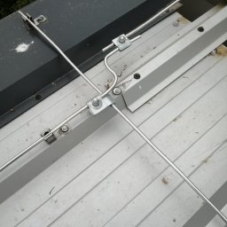 Elektro IT Sp.z.o.o - Metalowa instalacja odgromowa na szarej, blaszanej powierzchni dachu, widok z góry. Elementy mocujące z nakrętkami. Fragment czarnej skrzynki w lewym górnym rogu.