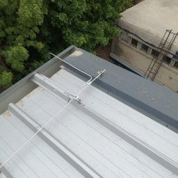 Elektro IT Sp.z.o.o - Metalowy dach z zamontowaną instalacją odgromową, widok z góry, z bliska. W tle zieleń drzew i budynek. Szare elementy konstrukcyjne.