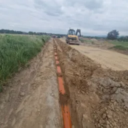 Wykonane wykopy pod pomarańczowe rury kanalizacyjne na budowie, w tle koparka Mecalac i pochmurne niebo. Widok z perspektywy ziemi.