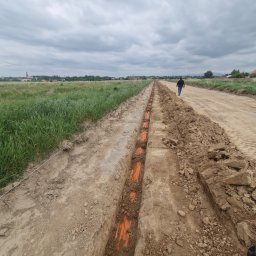 Elektro IT Sp.z.o.o - Wykop z pomarańczowymi rurami kablowymi ułożonymi w ziemi, widok z góry na długi rów, w tle spacerujący mężczyzna i krajobraz z wiatrakami.