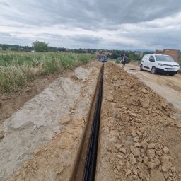 Elektro IT Sp.z.o.o - Wykop z ułożonymi rurami kablowymi na tle krajobrazu z zabudowaniami i białym samochodem dostawczym. Widok z góry na wykop.