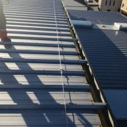 Elektro IT Sp.z.o.o - Metalowy dach z zamontowaną instalacją odgromową, widok z góry, fragment osoby w kadrze. Elementy mocujące widoczne wzdłuż dachu.