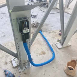 Montaż instalacji elektrycznej: metalowa konstrukcja z puszką i niebieską rurą karbowaną. Widoczne narzędzia i elementy montażowe na surowym podłożu.