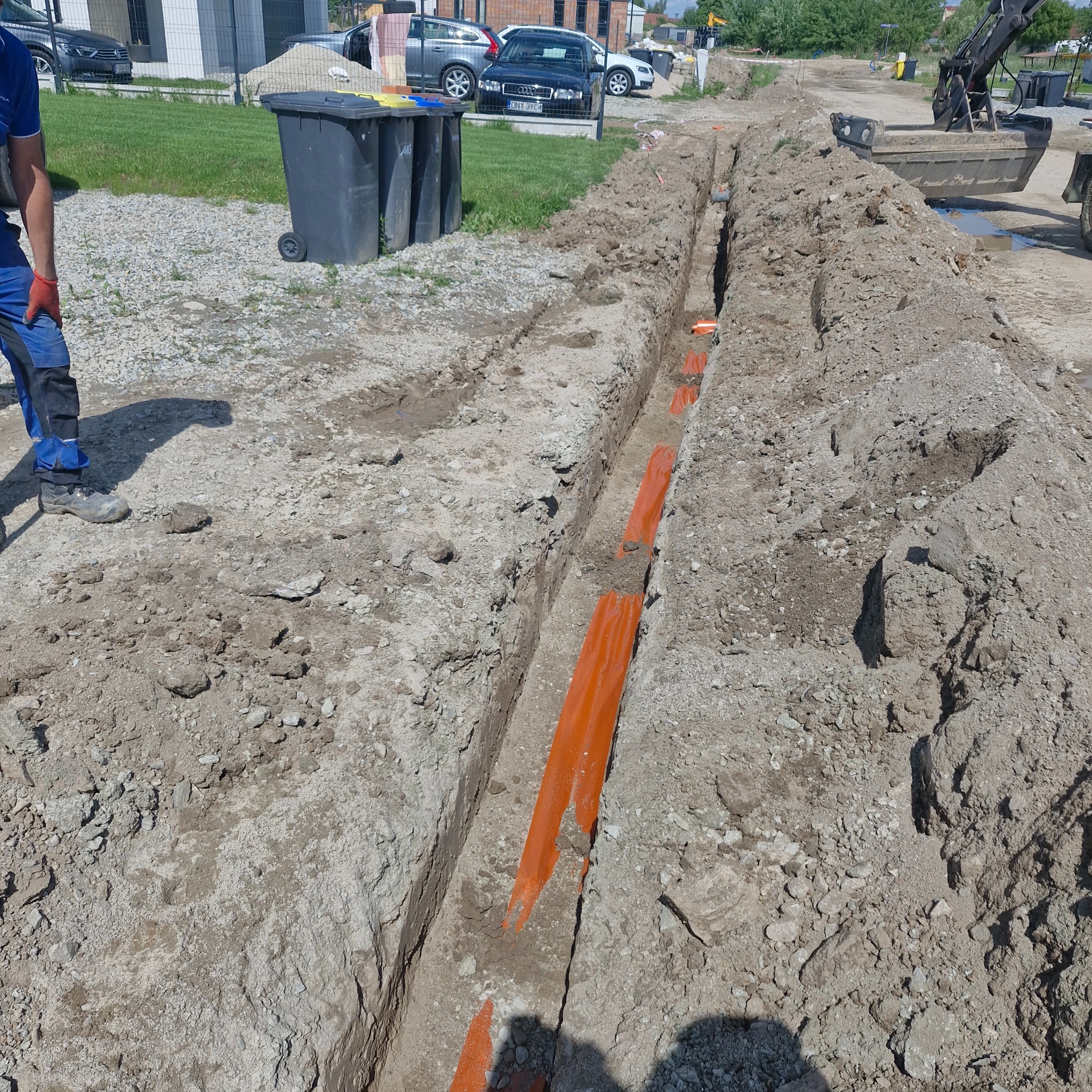 Wykop z pomarańczowymi rurami kablowymi, prawdopodobnie pod instalacje elektryczne, widok z góry, Opole. Prace ziemne w toku, koparka w tle.
