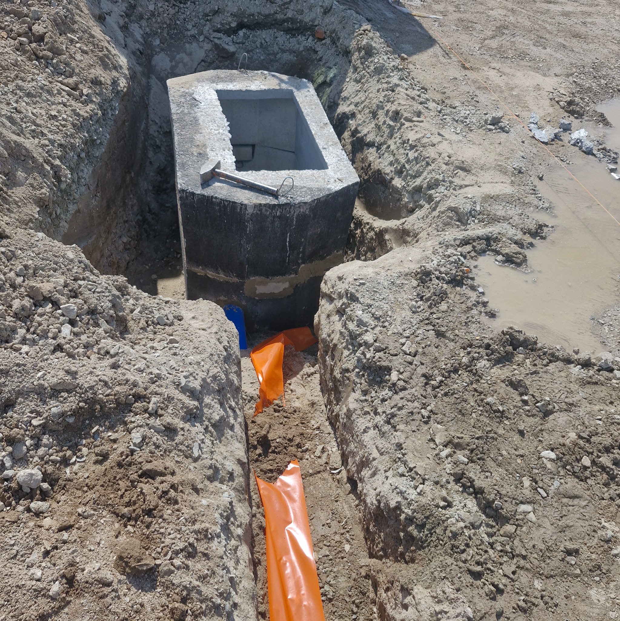 Betonowa studzienka w wykopie, z widocznym młotkiem na krawędzi i pomarańczową folią zabezpieczającą, sugerująca etap prac przy instalacji podziemnej.