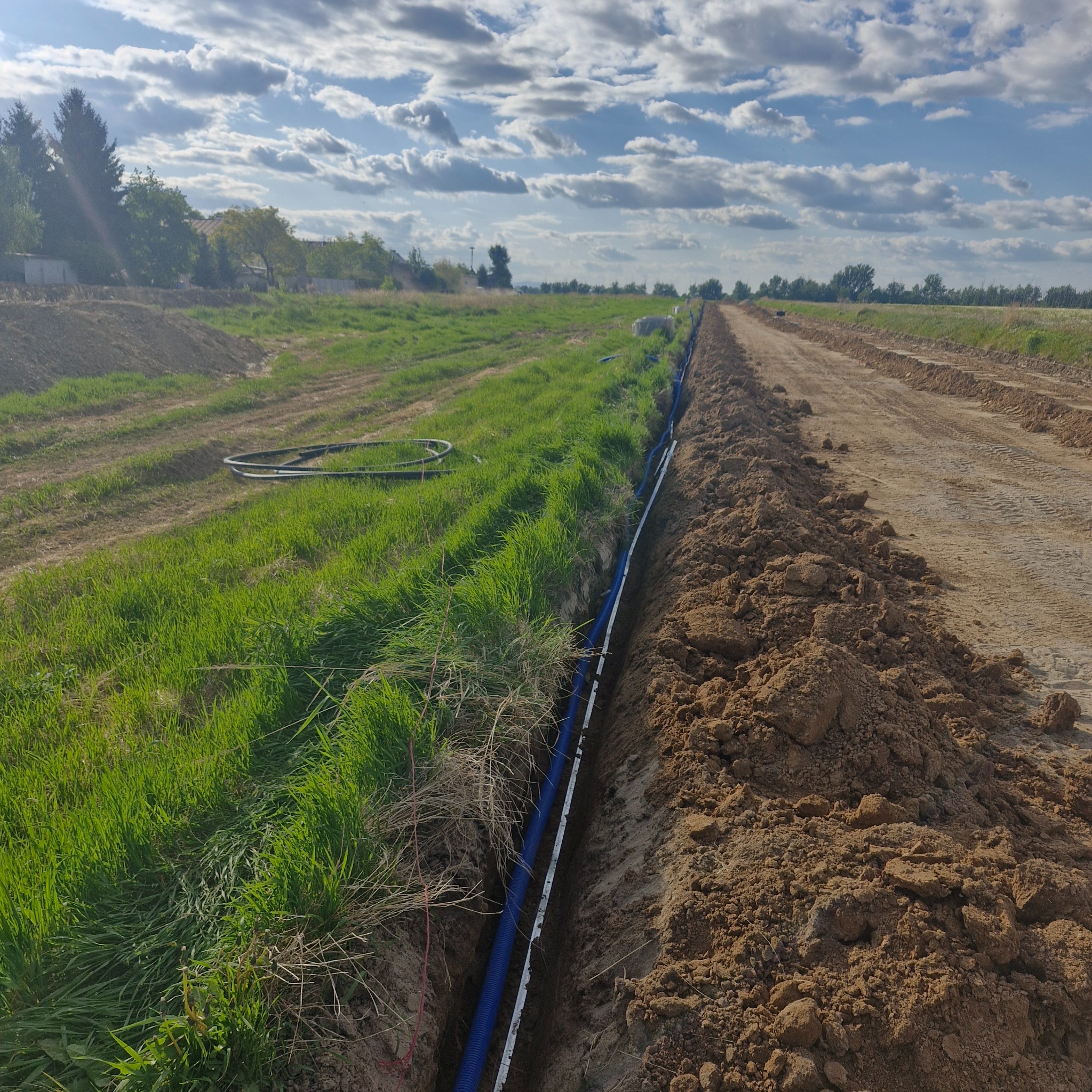 Błękitne rury kablowe ułożone w wykopie ziemnym na tle zielonej trawy i błękitnego nieba z chmurami. Instalacja kablowa w terenie otwartym.