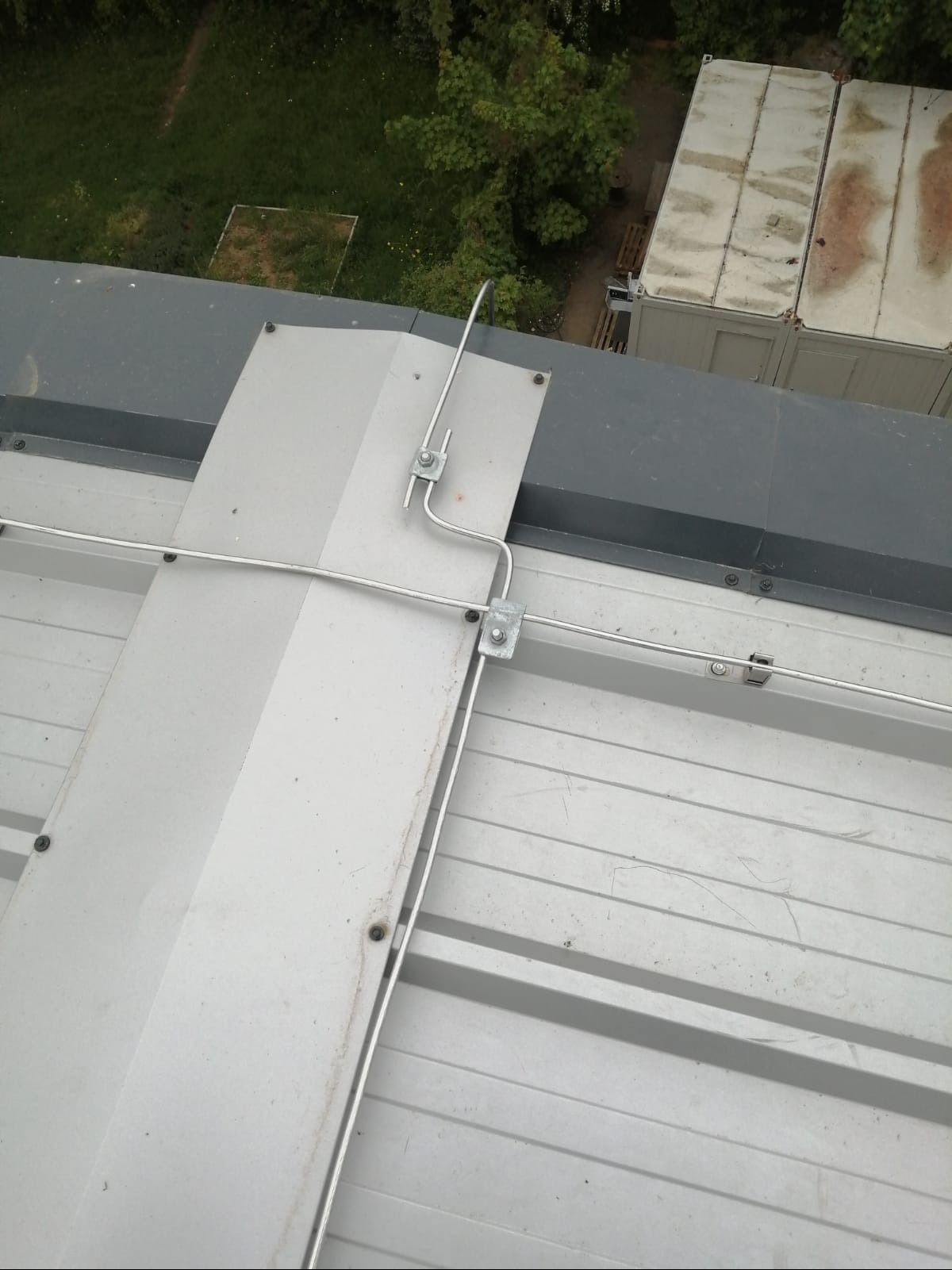 Metalowa instalacja odgromowa na dachu budynku z blachy trapezowej, widok z góry. Elementy połączone śrubami, tło stanowią kontenery i zieleń.