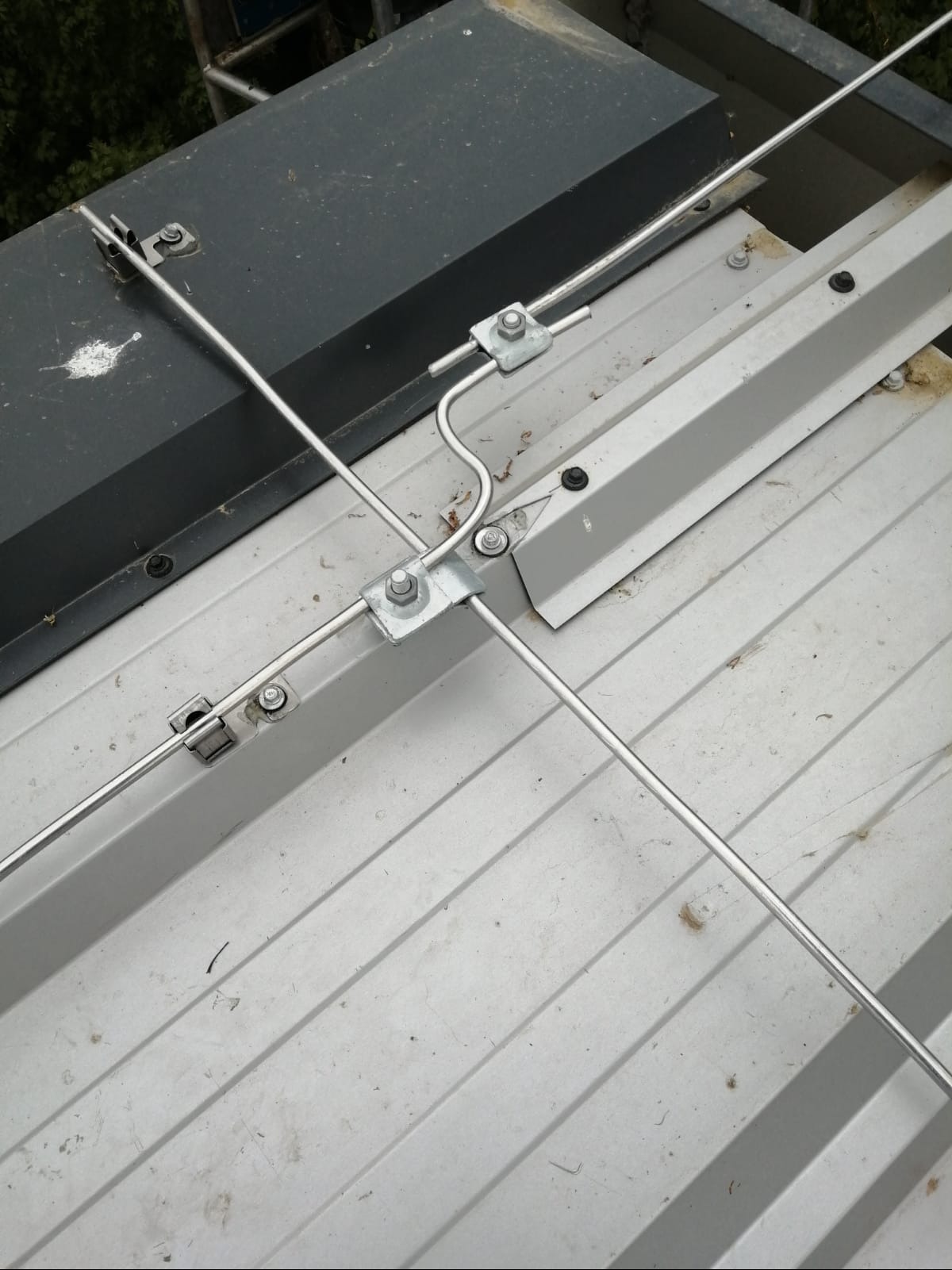 Metalowa instalacja odgromowa na szarej, blaszanej powierzchni dachu, widok z góry. Elementy mocujące z nakrętkami. Fragment czarnej skrzynki w lewym górnym rogu.