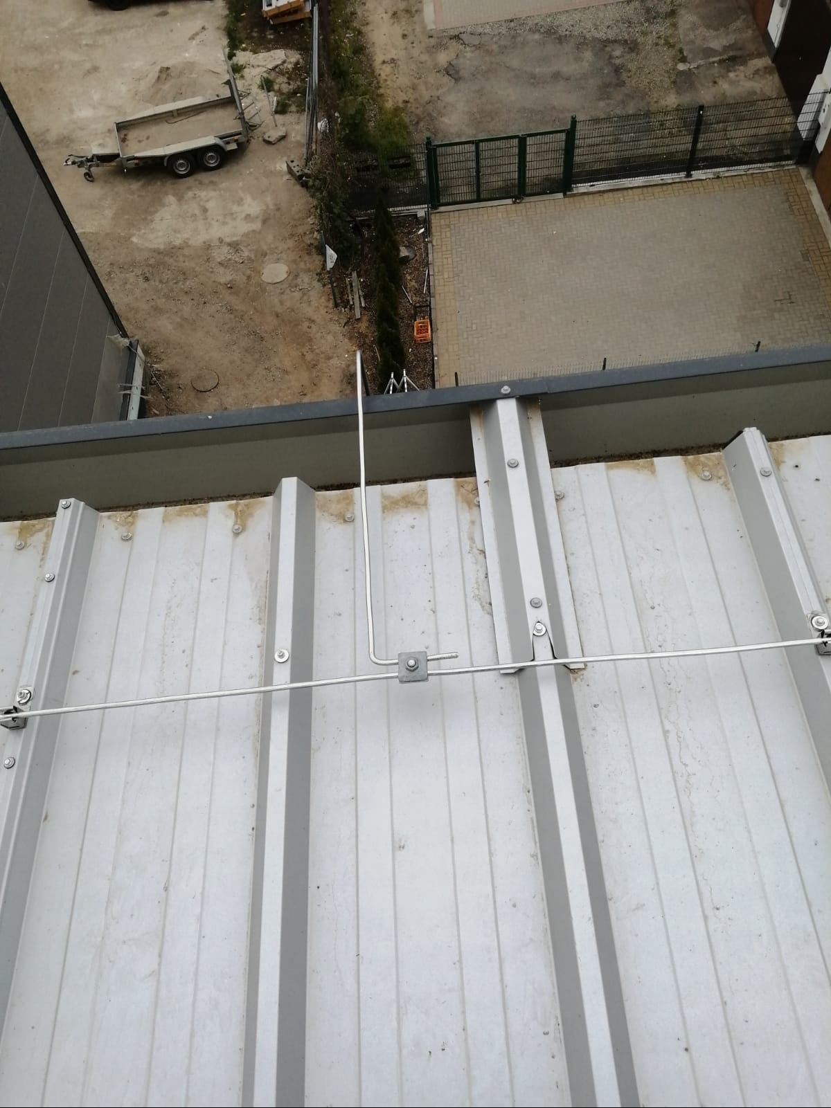 Metalowa instalacja odgromowa na dachu z blachy trapezowej, widok z góry, z fragmentem otoczenia budynku w tle.