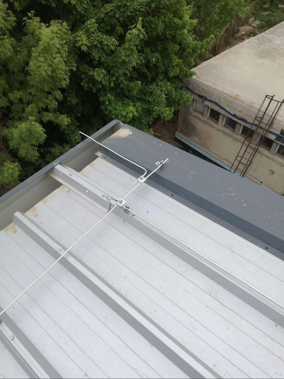 Metalowy dach z zamontowaną instalacją odgromową, widok z góry, z bliska. W tle zieleń drzew i budynek. Szare elementy konstrukcyjne.