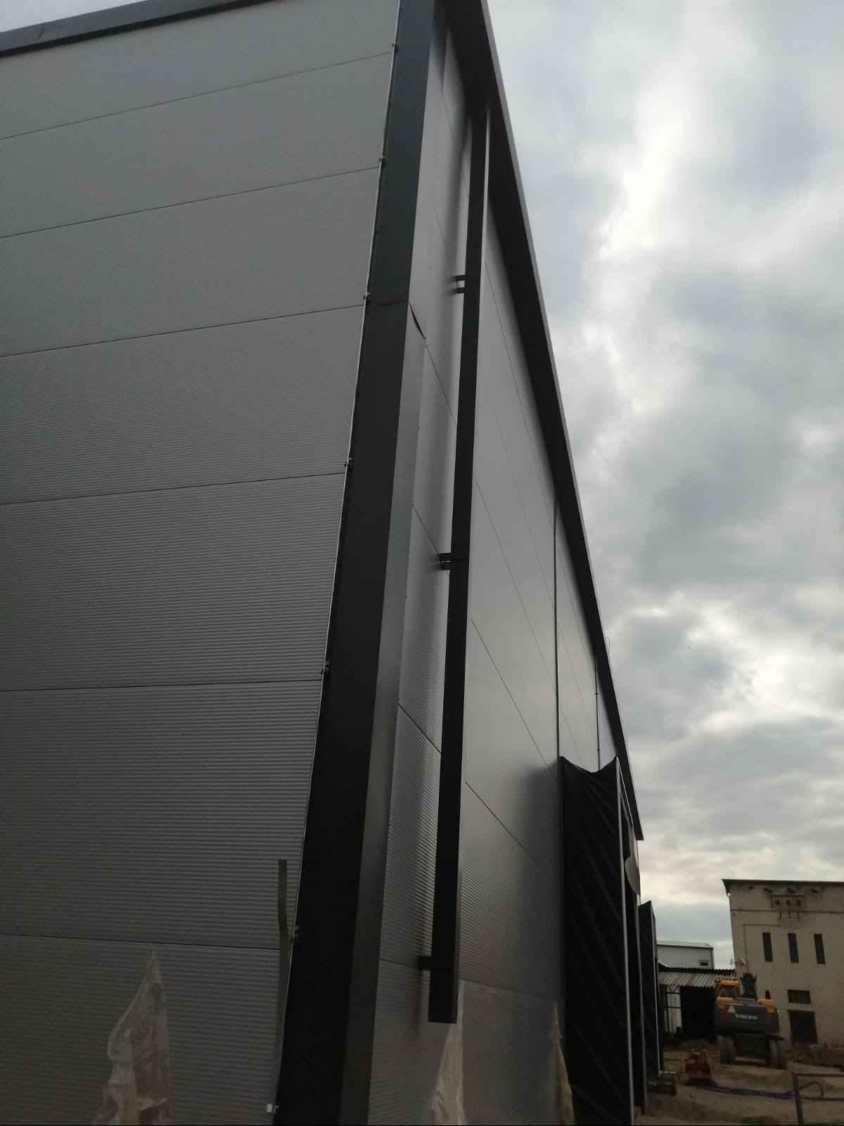 Elewacja hali przemysłowej z szarej blachy trapezowej, widok z dołu pod kątem, z elementami konstrukcyjnymi w kolorze grafitowym i pochmurnym niebem w tle.