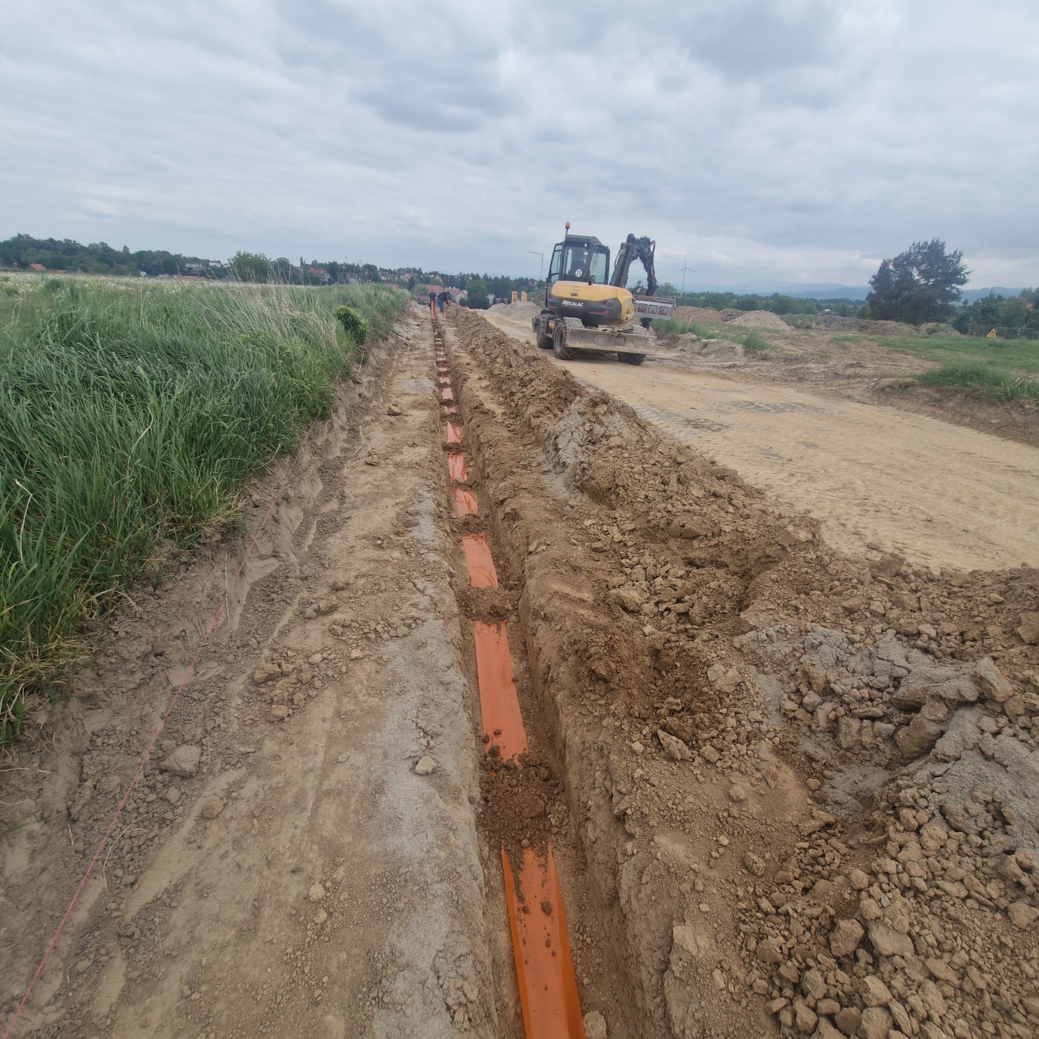 Wykonane wykopy pod pomarańczowe rury kanalizacyjne na budowie, w tle koparka Mecalac i pochmurne niebo. Widok z perspektywy ziemi.