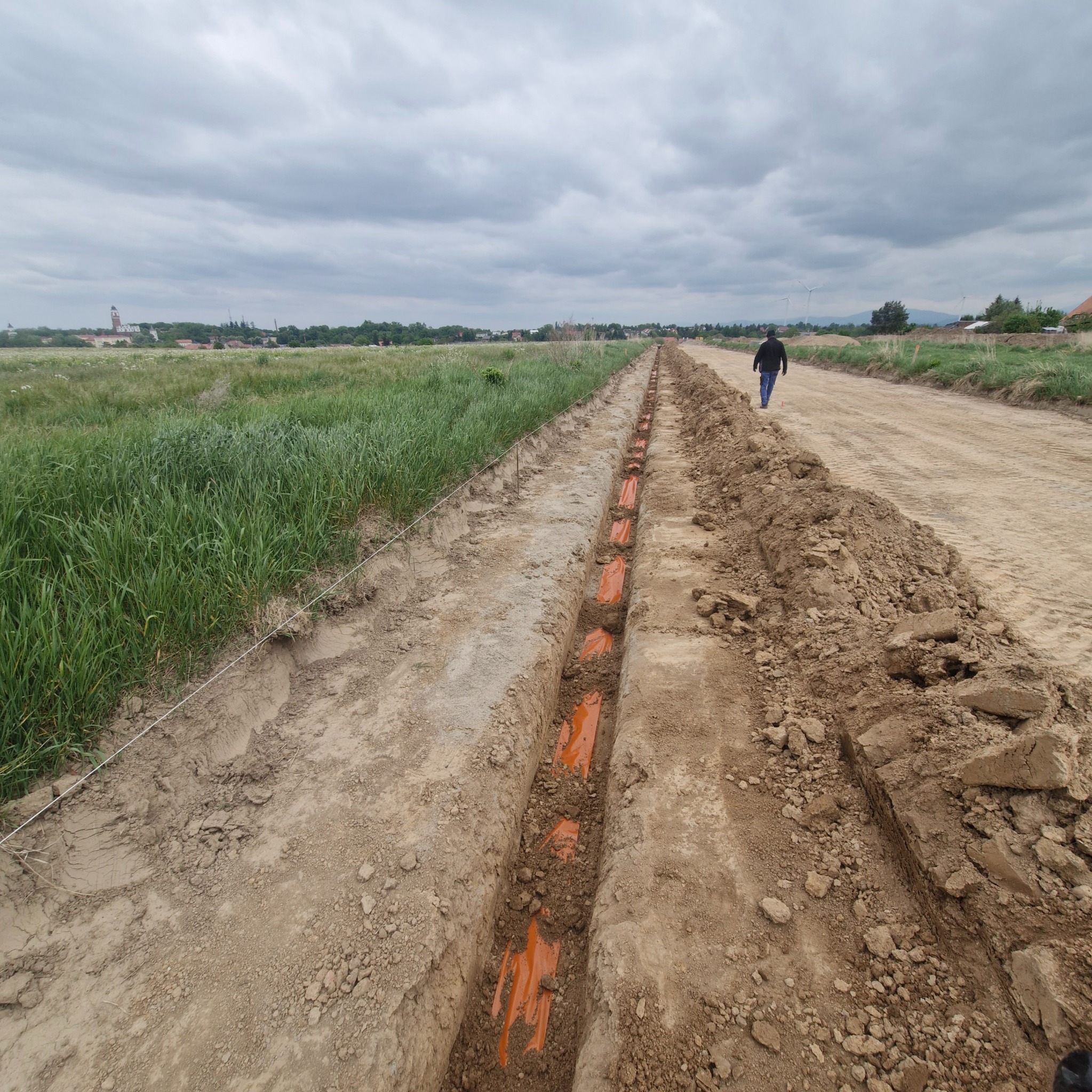 Wykop z pomarańczowymi rurami kablowymi ułożonymi w ziemi, widok z góry na długi rów, w tle spacerujący mężczyzna i krajobraz z wiatrakami.