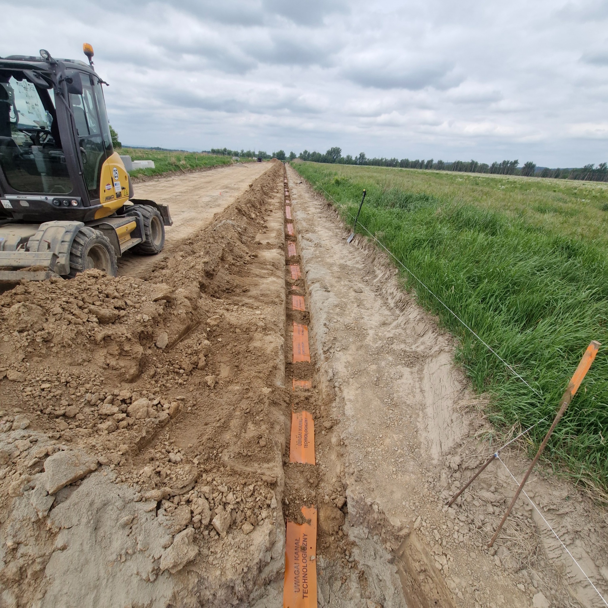 Wykop z ułożonymi pomarańczowymi rurami kablowymi w ziemi, widoczny fragment koparki i teren zielony. Prace ziemne przy instalacji kablowej.