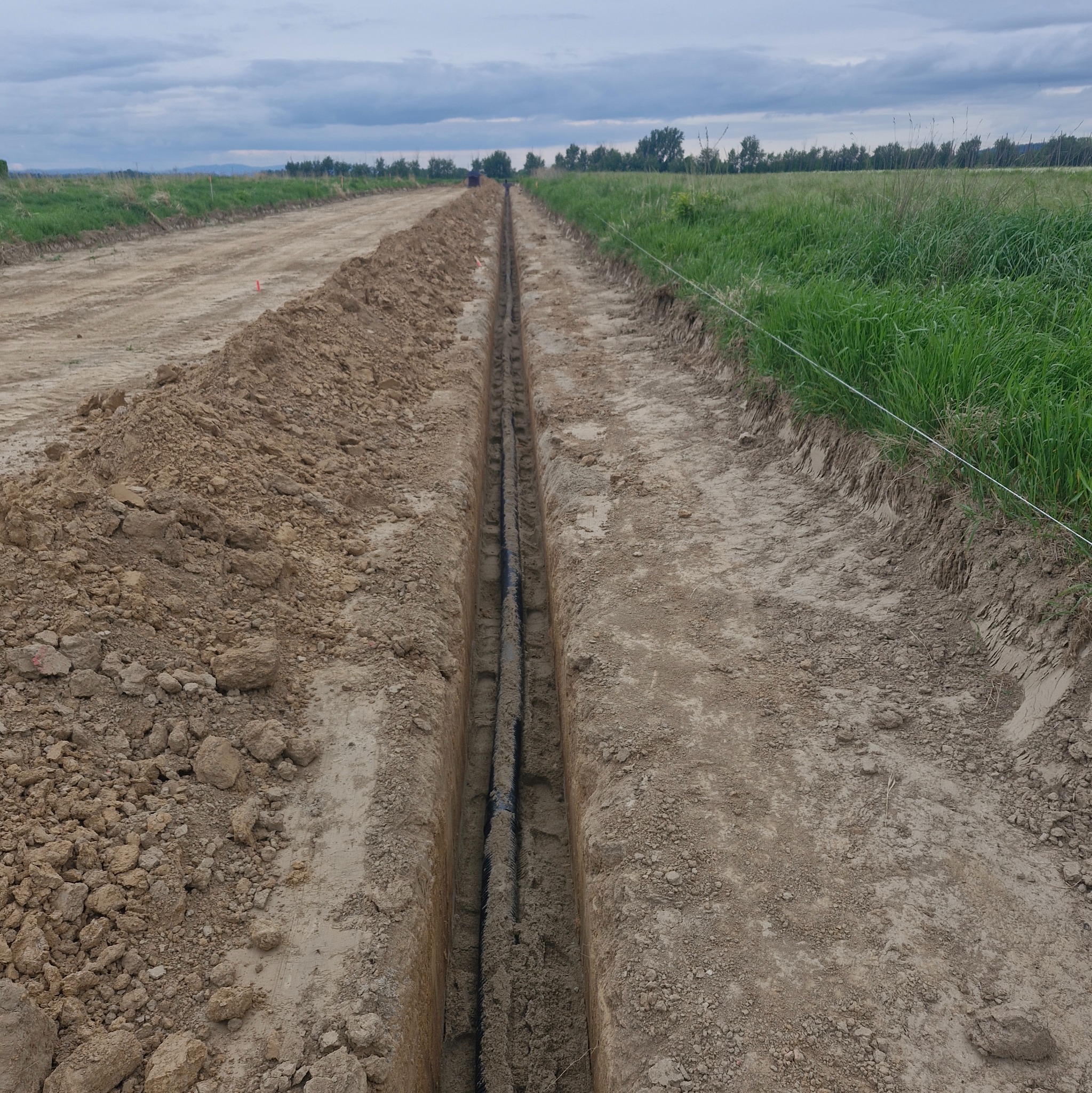 Wykop w ziemi z ułożonymi rurami, prawdopodobnie pod przyłącze elektryczne. Widoczne ślady po koparce i urobiona ziemia po bokach wykopu. Trawa i drzewa w tle.