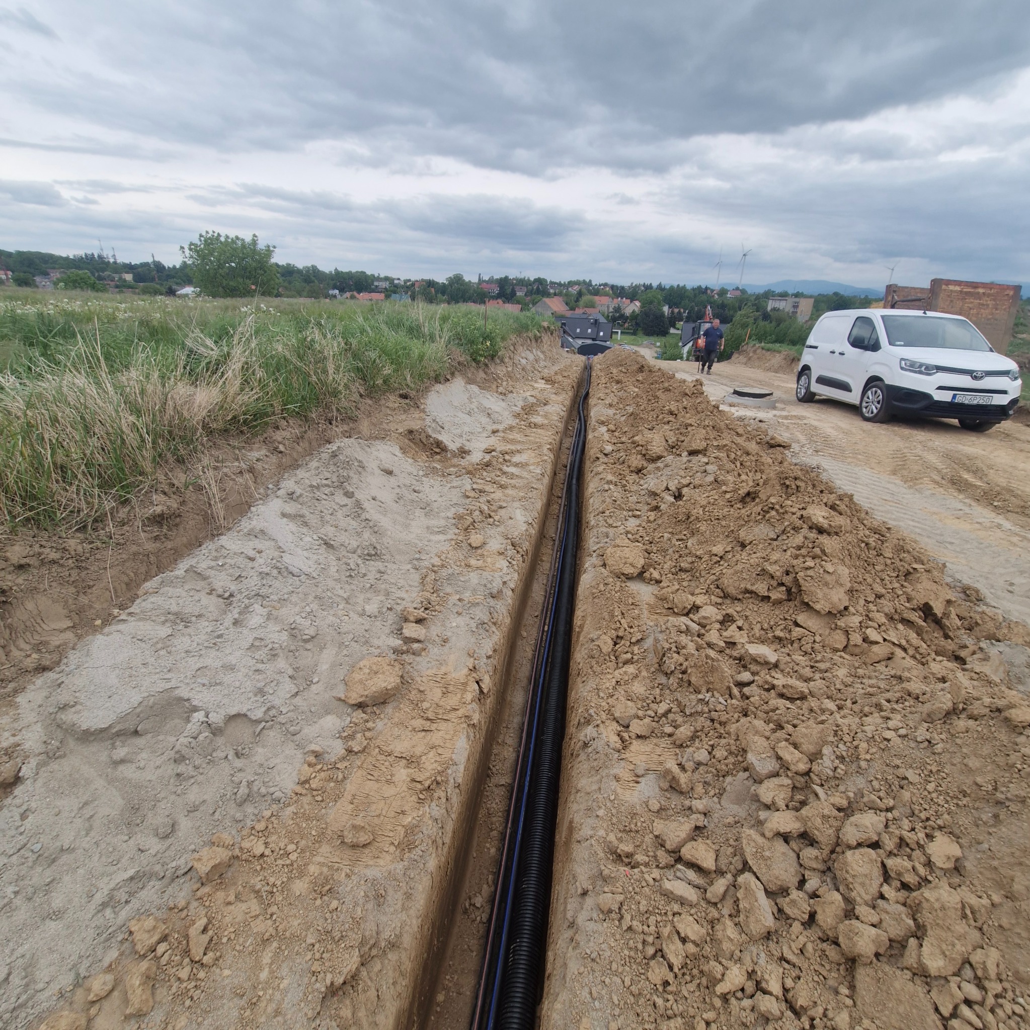 Wykop z ułożonymi rurami kablowymi na tle krajobrazu z zabudowaniami i białym samochodem dostawczym. Widok z góry na wykop.