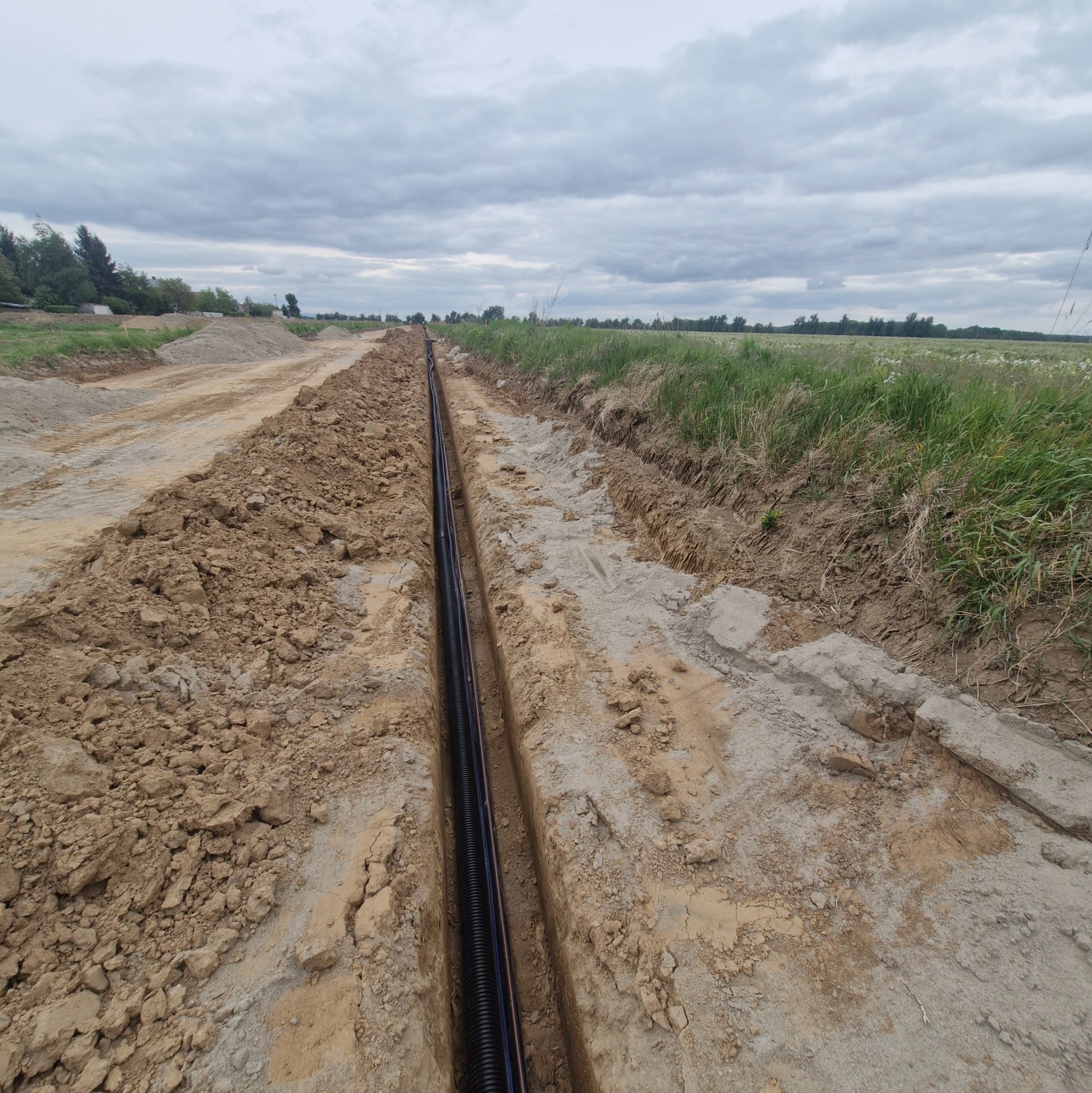 Rów z czarnym karbowanym przewodem w ziemi na tle pochmurnego nieba i łąki. Przygotowanie do ułożenia kabli elektrycznych.