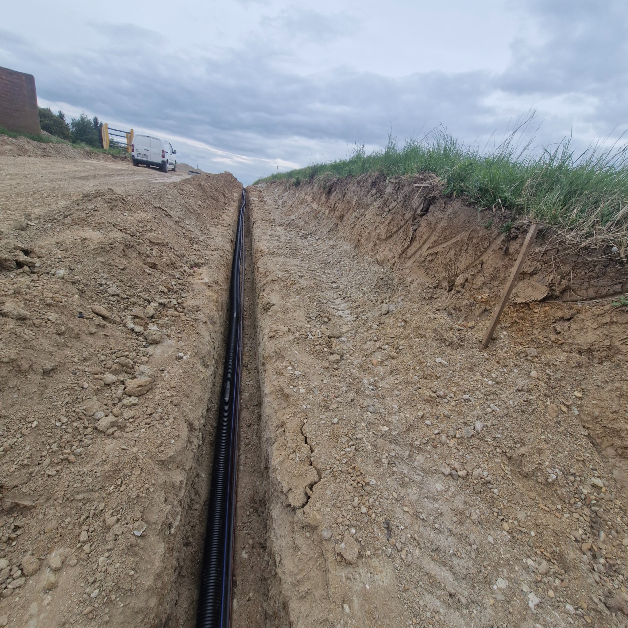 Wykop z ułożonymi rurami kablowymi w ziemi, widok z góry. Na górze trawa, w tle zaparkowany samochód dostawczy.