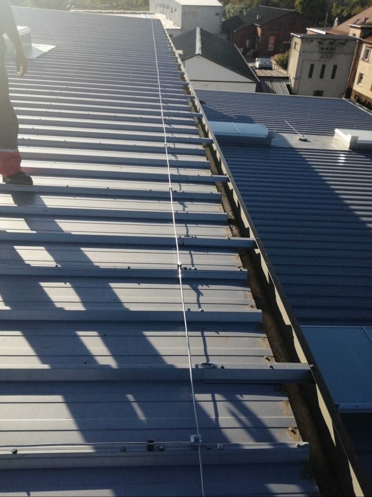 Metalowy dach z zamontowaną instalacją odgromową, widok z góry, fragment osoby w kadrze. Elementy mocujące widoczne wzdłuż dachu.