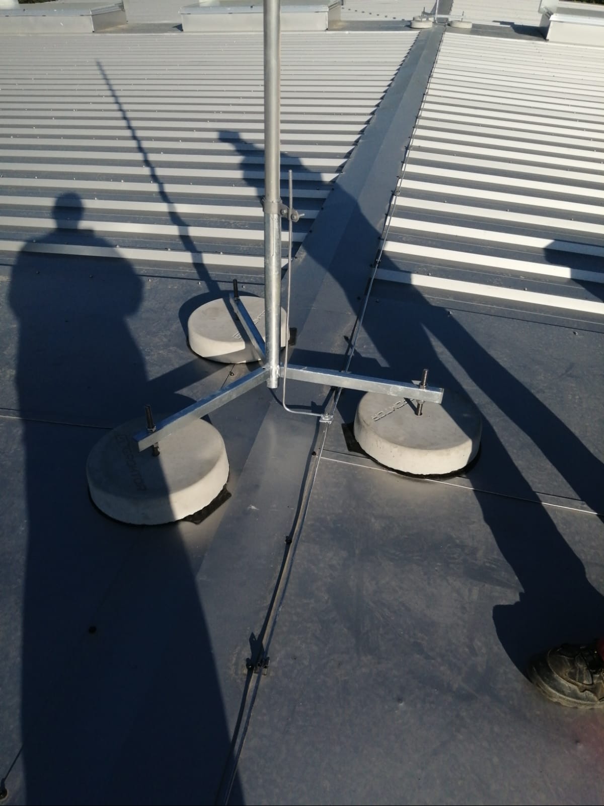 Szara instalacja odgromowa z trzema betonowymi podstawami na metalowym dachu. Cień osoby widoczny w lewym rogu. Metalowe elementy mocujące.