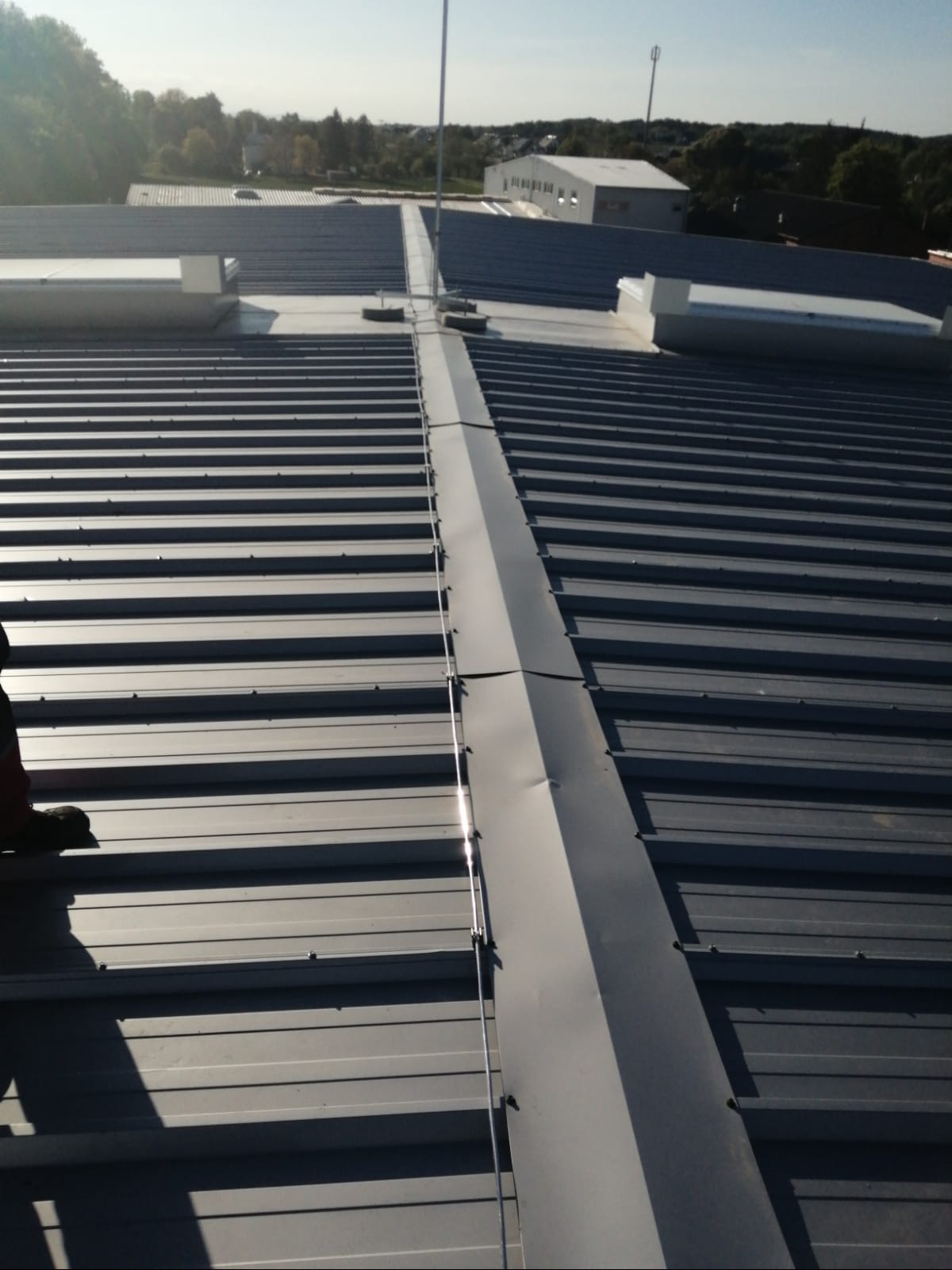 Szary blaszany dach budynku z zamontowaną instalacją odgromową. Widoczne mocowania i przewody. W tle zabudowania i drzewa.