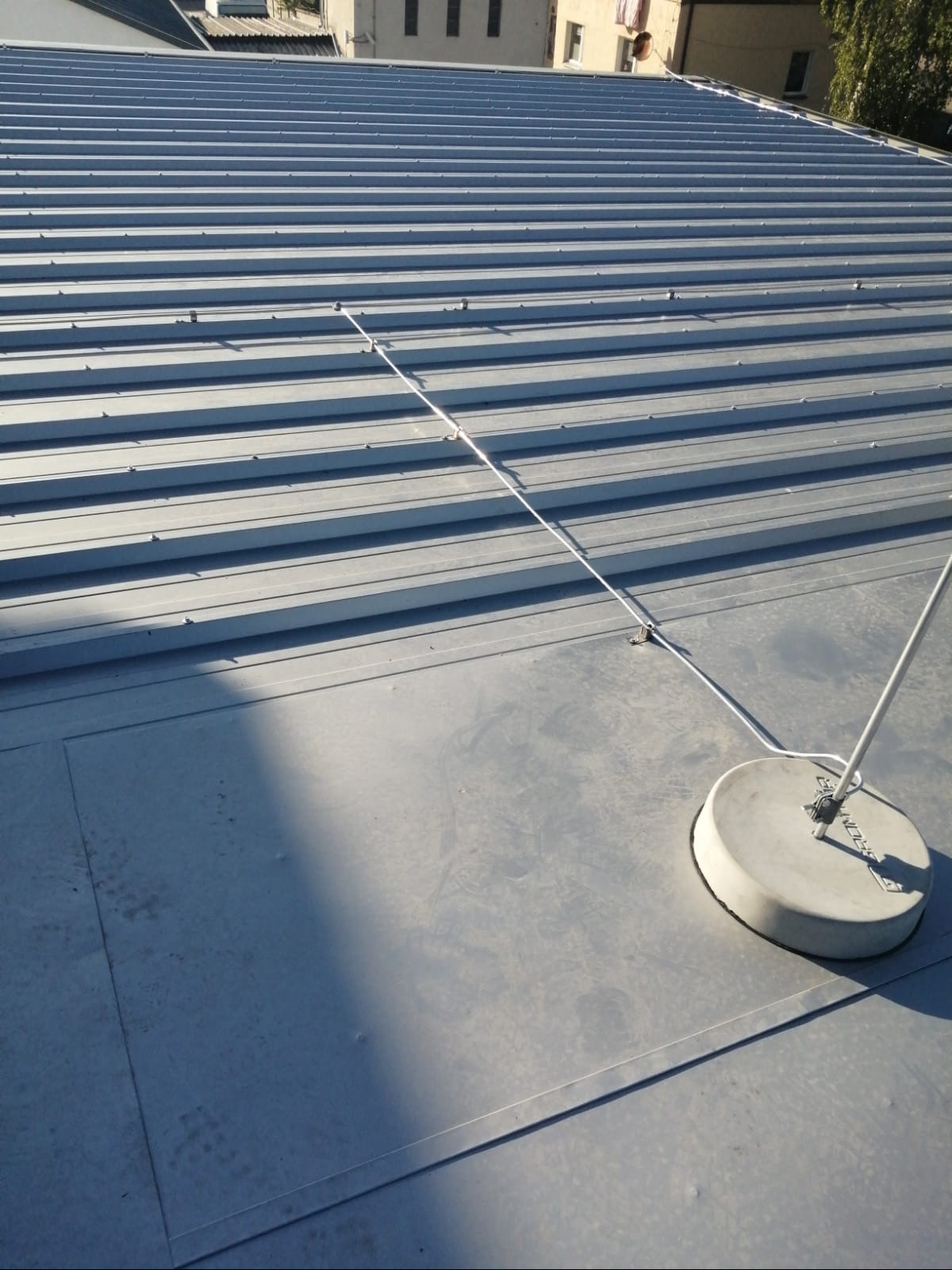 Szary dach z blachy trapezowej z zamontowaną instalacją odgromową. Widoczne przewody i elementy mocujące. W tle budynki.