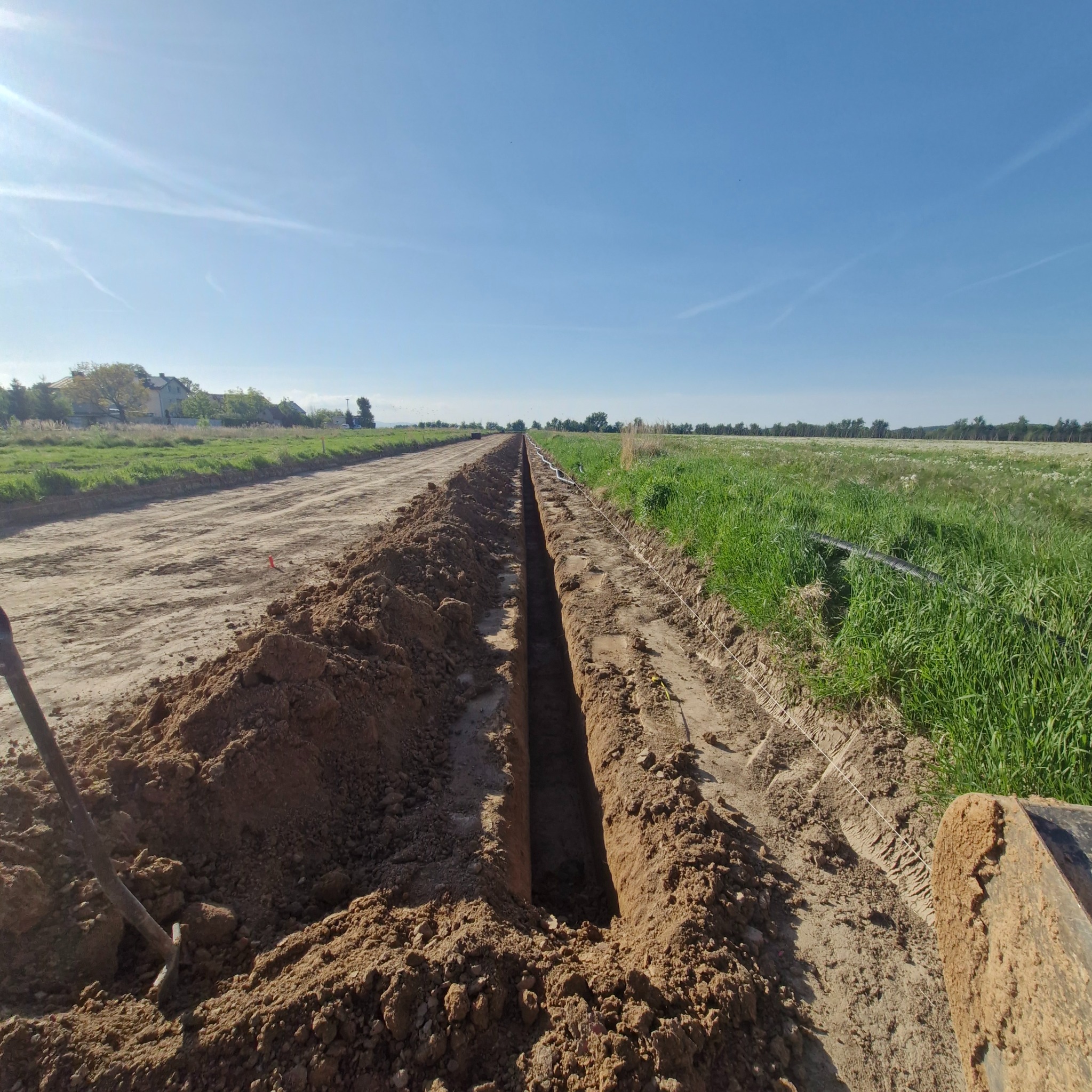 Wykopany rów w ziemi, prawdopodobnie pod instalację elektryczną, z widoczną rurą w tle i narzędziami po lewej stronie. Praca w toku na tle zieleni i błękitnego nieba.