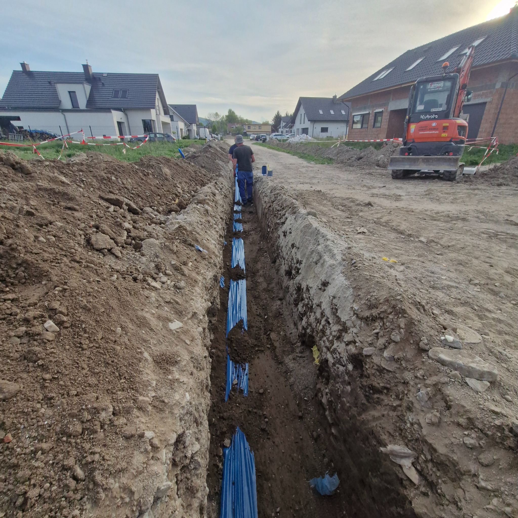 Wykop z niebieską rurą w ziemi na tle domów jednorodzinnych i koparki Kubota, sugerujący prace przyłączeniowe.