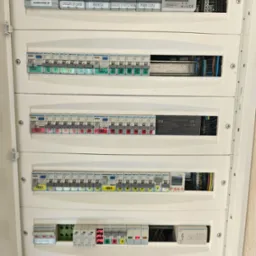 Rozdzielnica elektryczna z bezpiecznikami, opisana fazami i obwodami, widoczne elementy pomiarowe i zabezpieczenia.