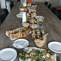 Młyn catering - Stół cateringowy z przekąskami: kanapki, sałatki, tortille. W tle rząd butelek Coca-Coli na szafce. Widok z góry, oświetlenie naturalne.