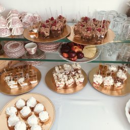 Młyn catering - Elegancki bufet deserowy z różnorodnymi słodkościami: tartaletki z bitą śmietaną, desery w kubeczkach z owocami, ułożone na złotych paterach.