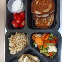 Młyn catering - Czarny pojemnik cateringowy z pancakes, sosem, truskawkami, kaszą i warzywami. Ryba z cytryną w dolnej przegródce. Zdrowy, zbilansowany posiłek.