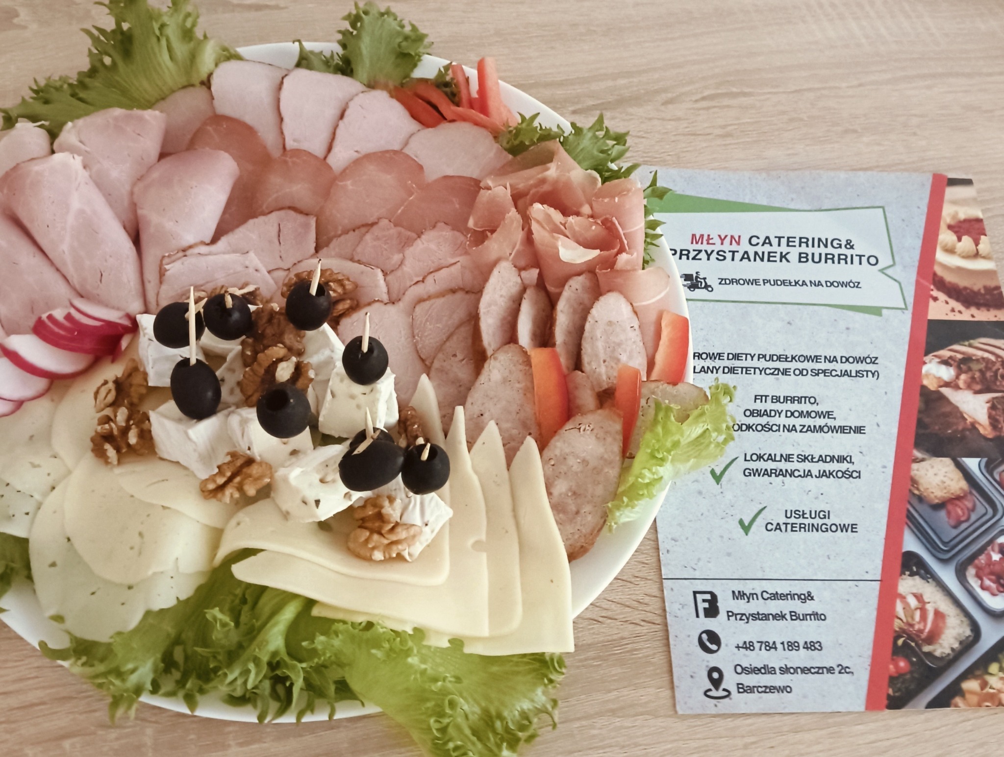 Dekoracyjna taca z wędlinami, serami i oliwkami, obok ulotka 'Młyn Catering & Przystanek Burrito', zdrowe pudełka na dowóz, lokalne składniki.