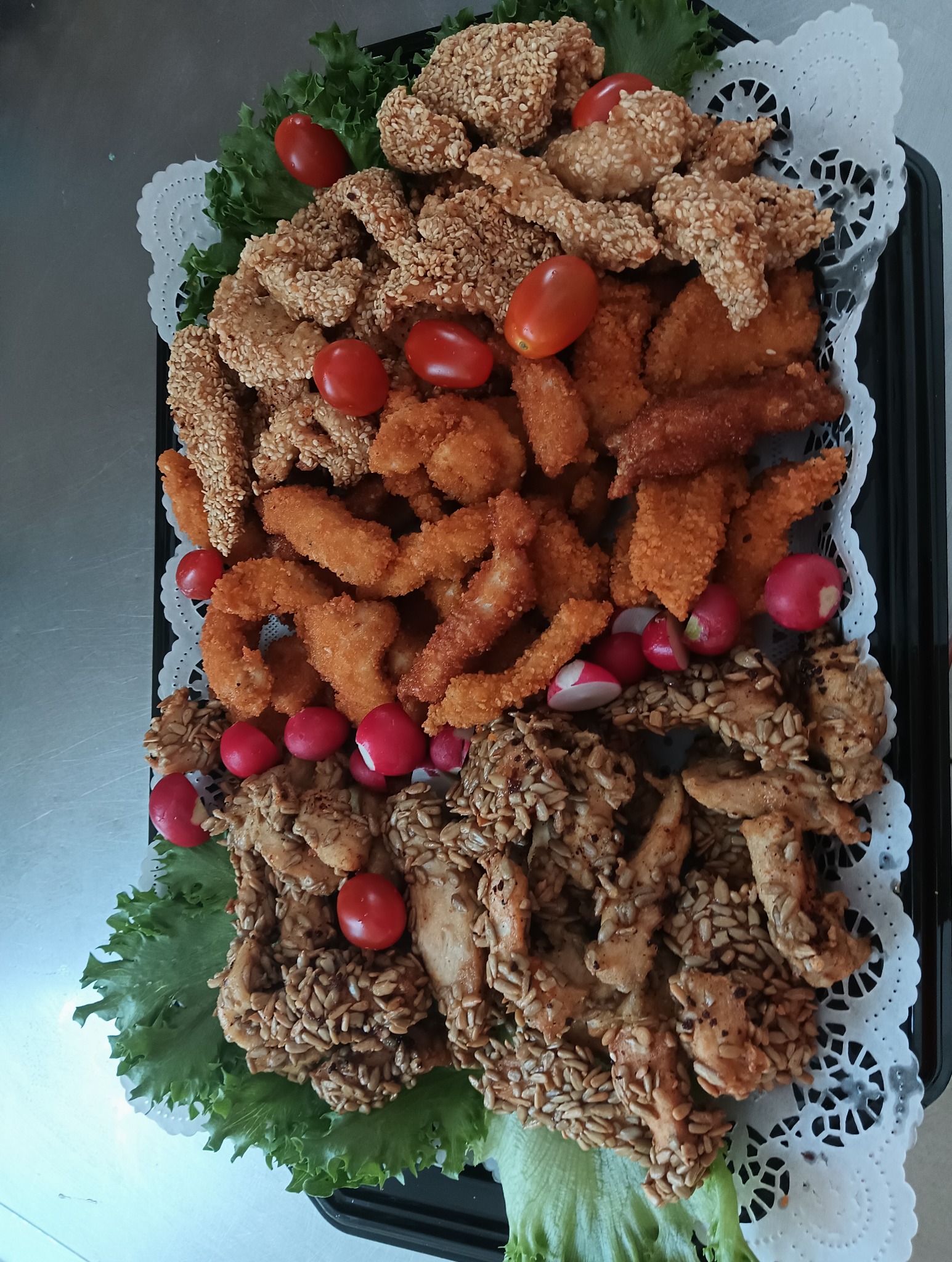 Czarna taca z różnorodnymi przekąskami: kurczak w sezamie i słoneczniku, nuggetsy, pomidorki cherry i rzodkiewki na dekoracyjnej serwetce.