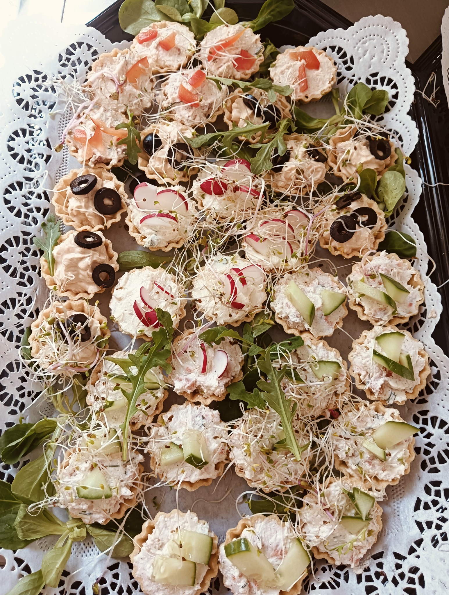 Kruche babeczki z kremowym nadzieniem i świeżymi dodatkami: rzodkiewką, ogórkiem, oliwkami i kiełkami, ułożone na ozdobnej serwetce.