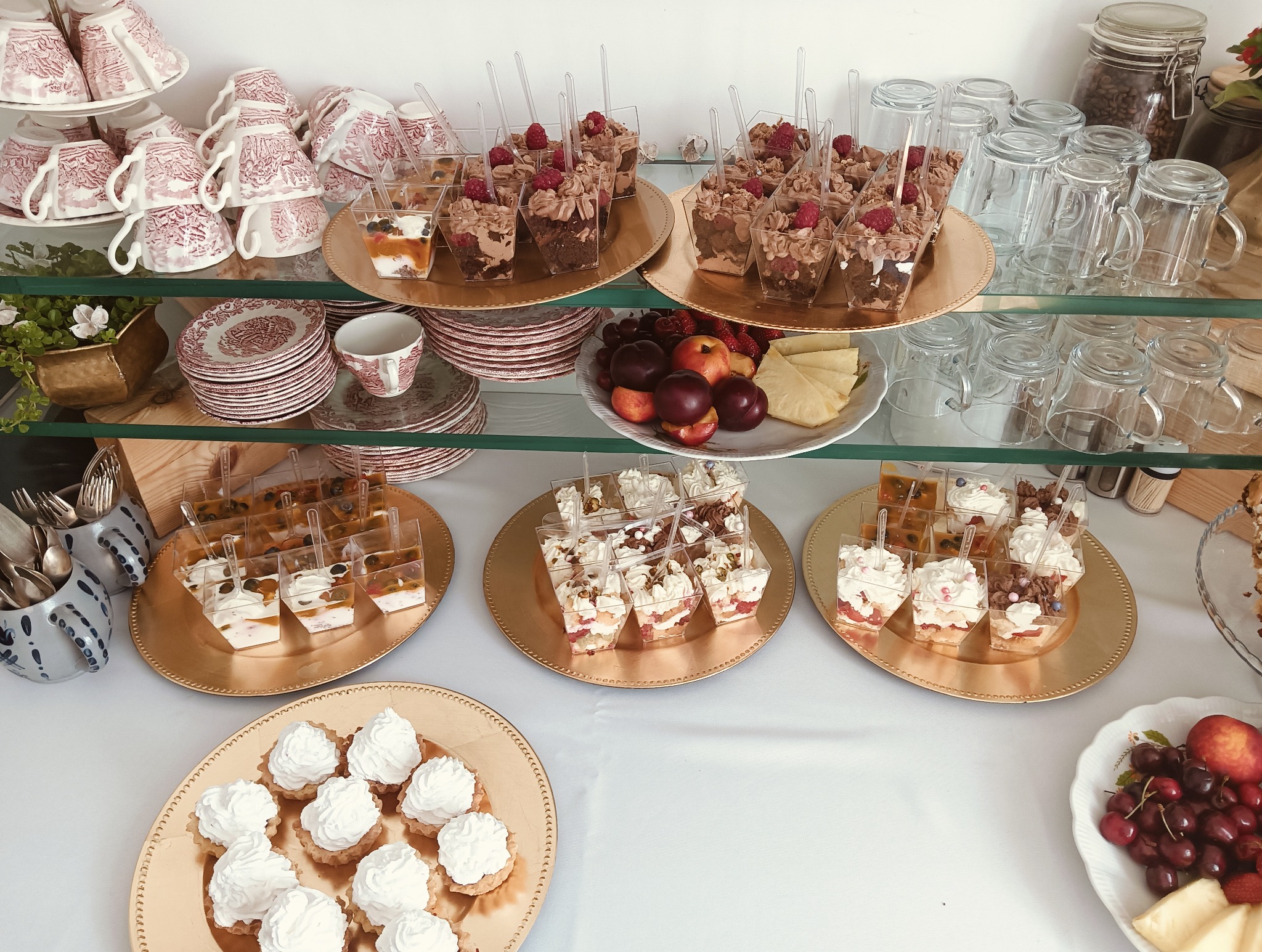 Bogaty wybór deserów w szklanych naczyniach na złotych paterach, uzupełniony owocami i elegancką zastawą, tworzący wykwintną kompozycję.