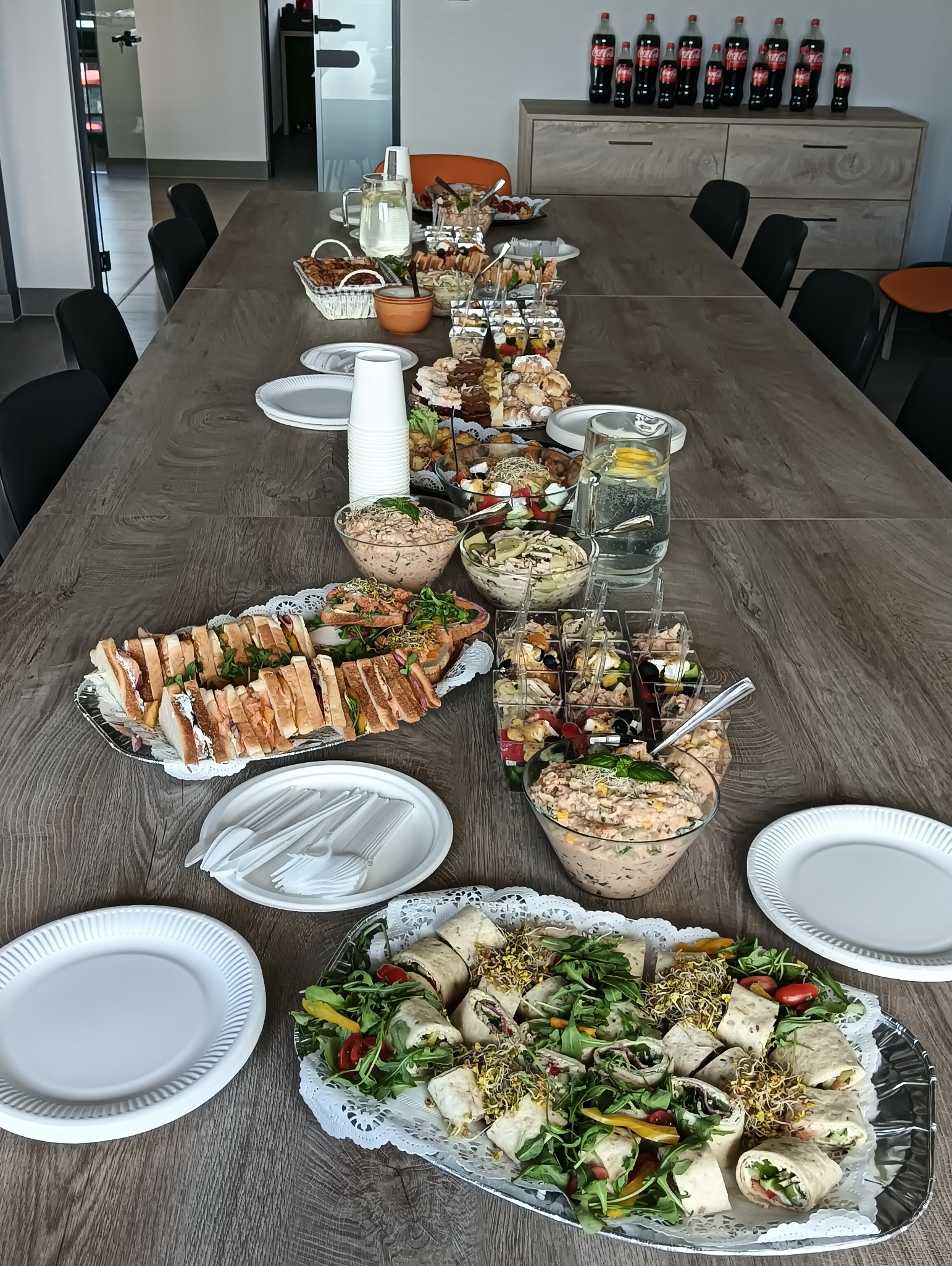 Stół cateringowy z przekąskami: kanapki, sałatki, tortille. W tle rząd butelek Coca-Coli na szafce. Widok z góry, oświetlenie naturalne.